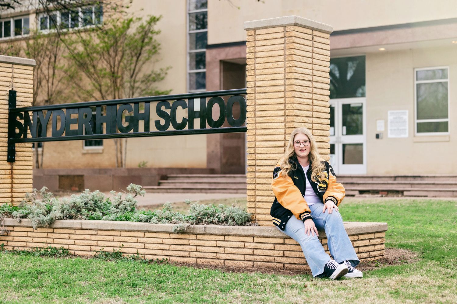 A student sits on grass outside a school building while studying and taking notes in a notebook.