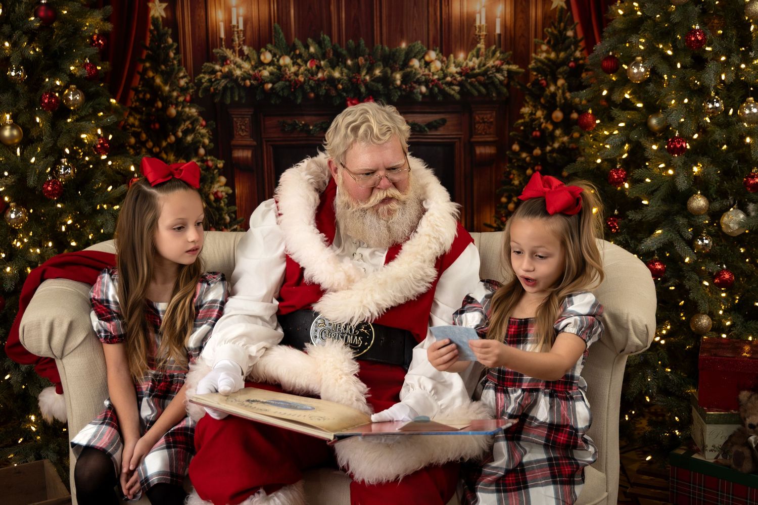 Two girls in plaid pajamas sit with Santa Claus reading a book beside decorated Christmas trees.