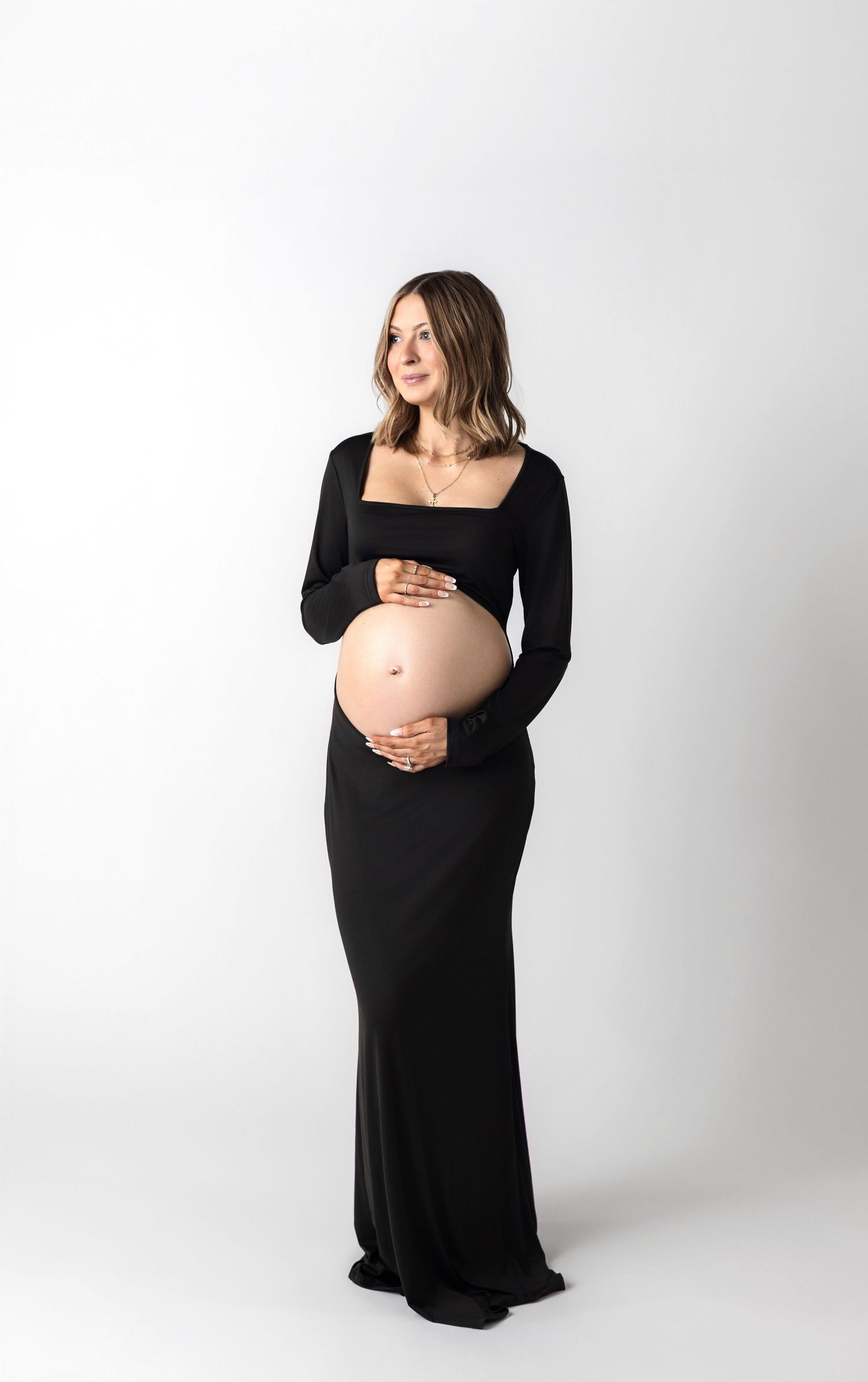 Expectant mother in long black dress poses against white backdrop in artistic maternity photoshoot series.