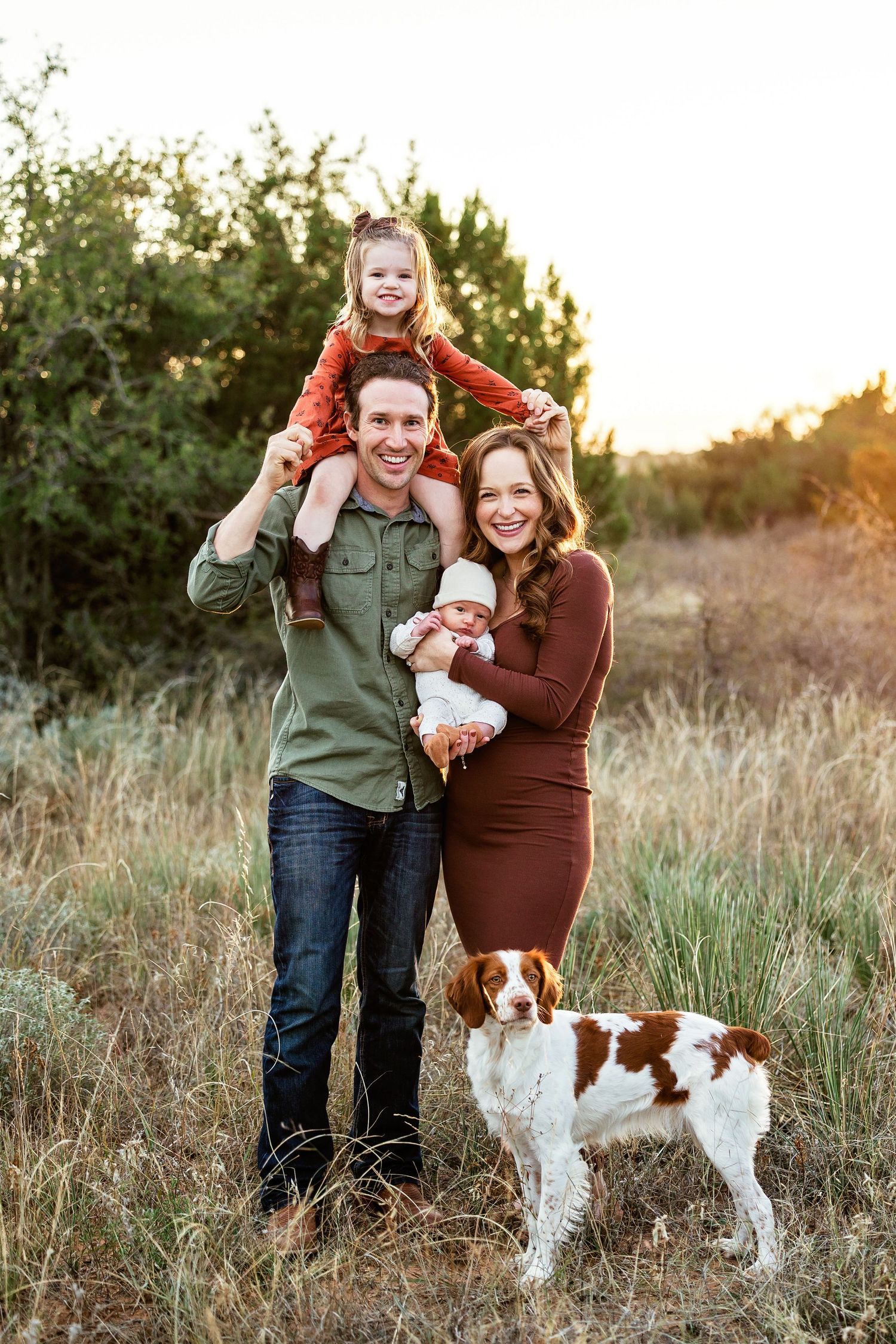 A couple poses playfully in a golden field at sunset with their brown and white dog.