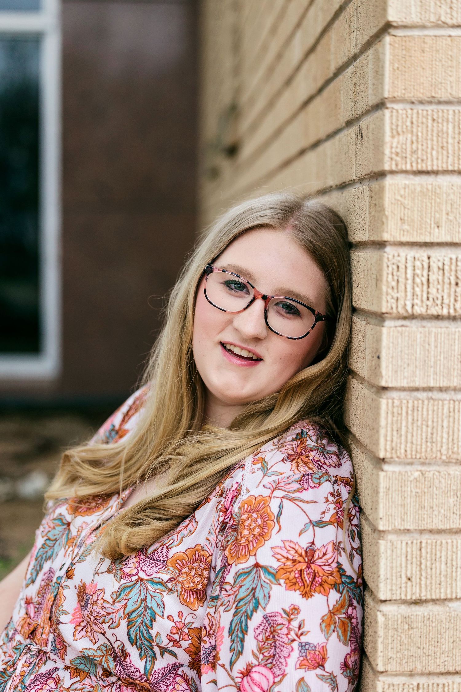 A person wearing glasses and a floral print top poses against a beige brick wall.