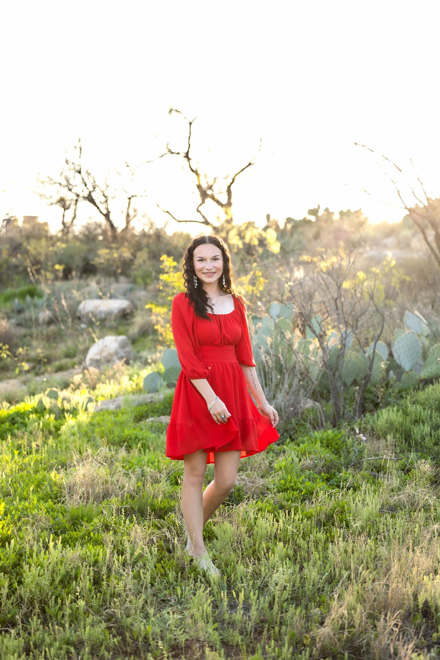A red dress flows gracefully in a golden sunlit meadow with wildflowers and natural foliage at sunset.