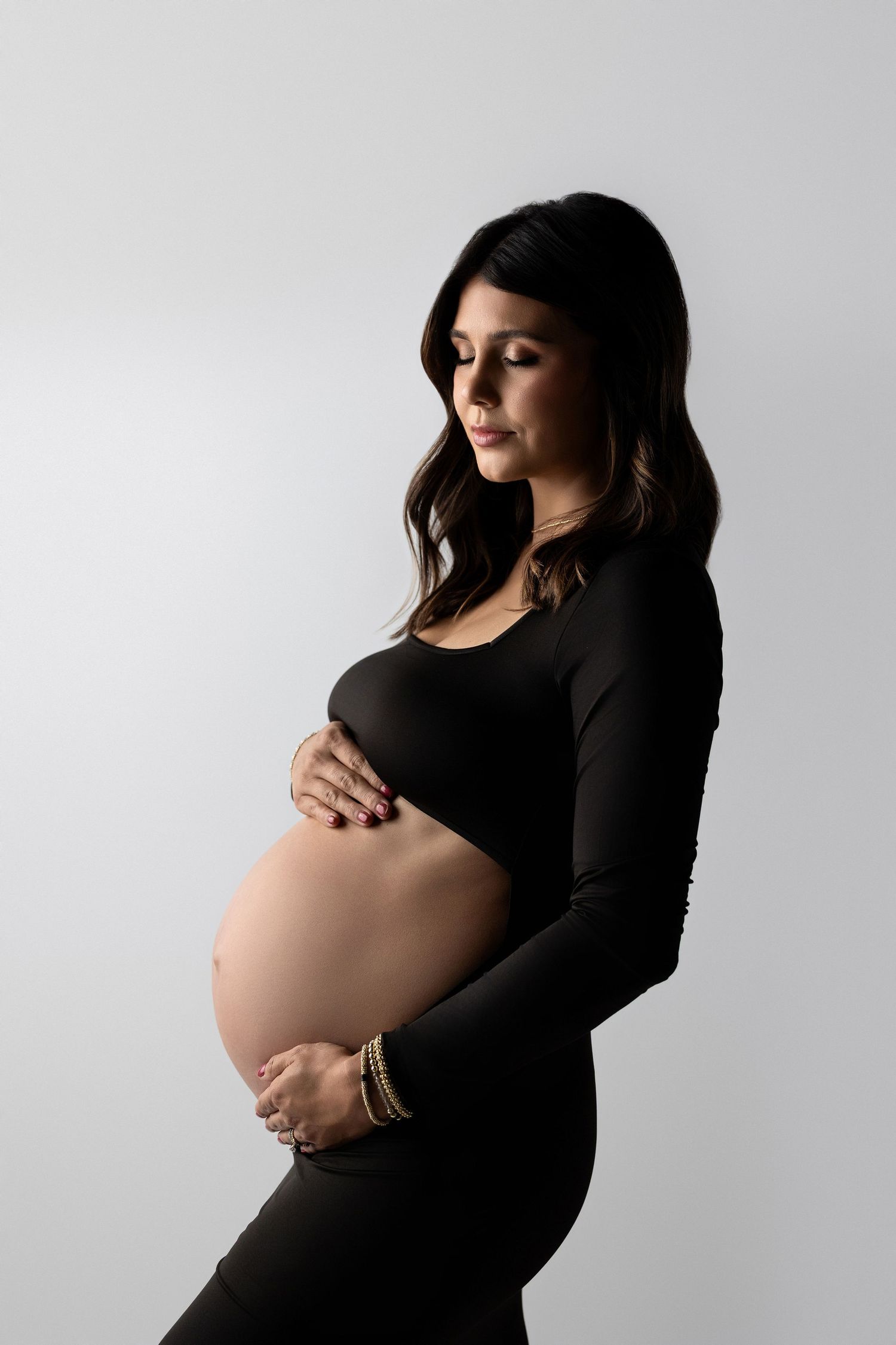 A silhouette style maternity photoshoot shows an expectant mother in a black dress against a light gray background.