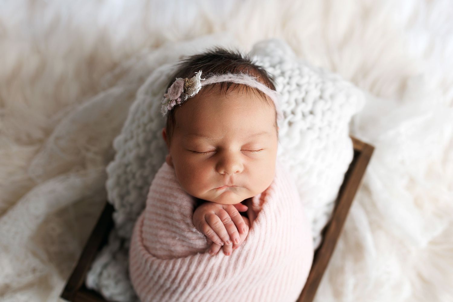 Sleeping newborn wrapped in soft pink swaddle wearing delicate white lace bonnet rests peacefully on cream blanket.