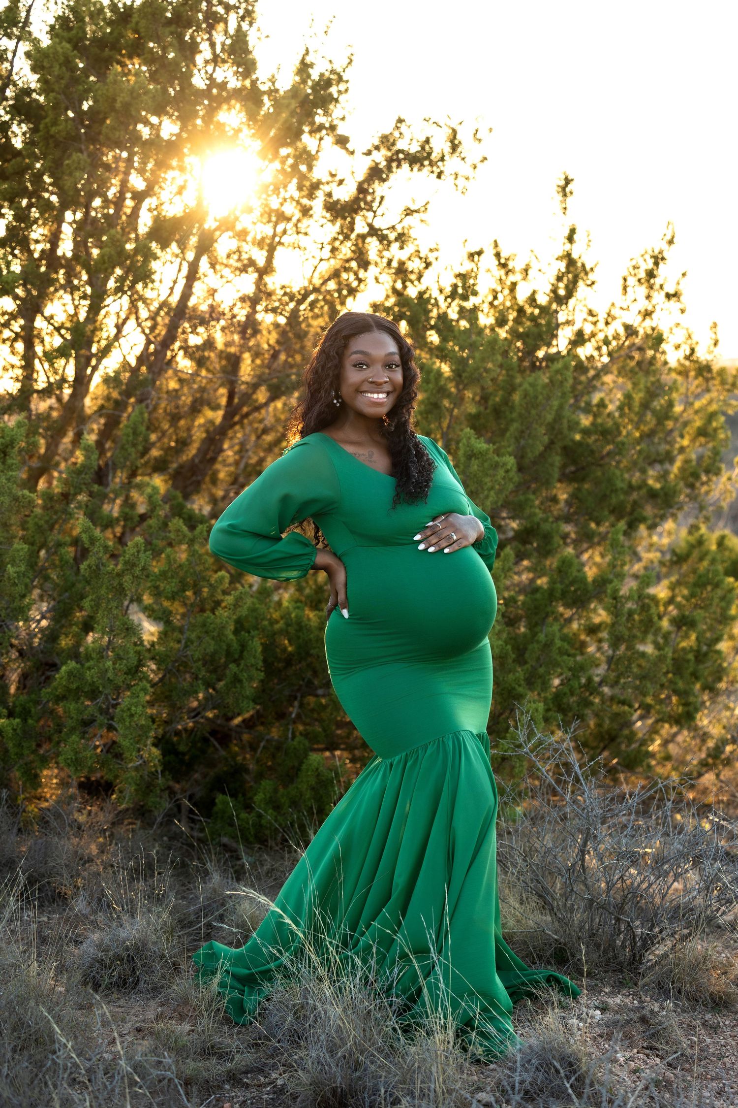 Woman in flowing emerald green dress poses gracefully in outdoor setting during golden hour sunset.