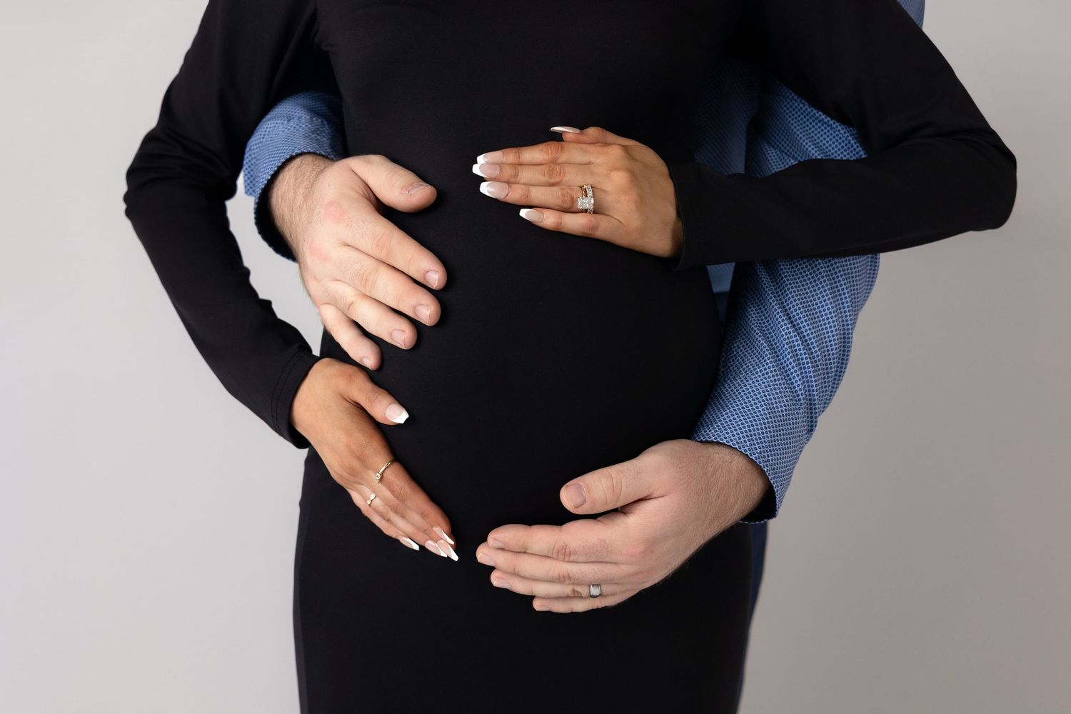 A pregnant person wearing a black dress and denim jacket poses against a gray backdrop in a maternity photoshoot.