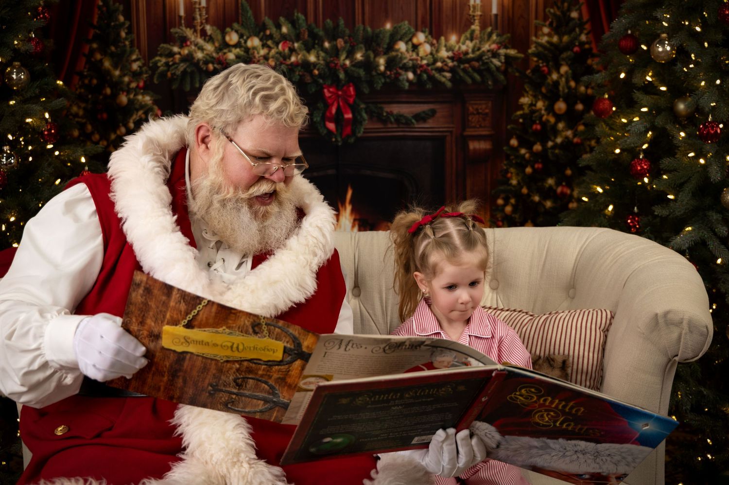 Santa Claus reads a Christmas storybook with a young girl by a decorated fireplace and Christmas tree.