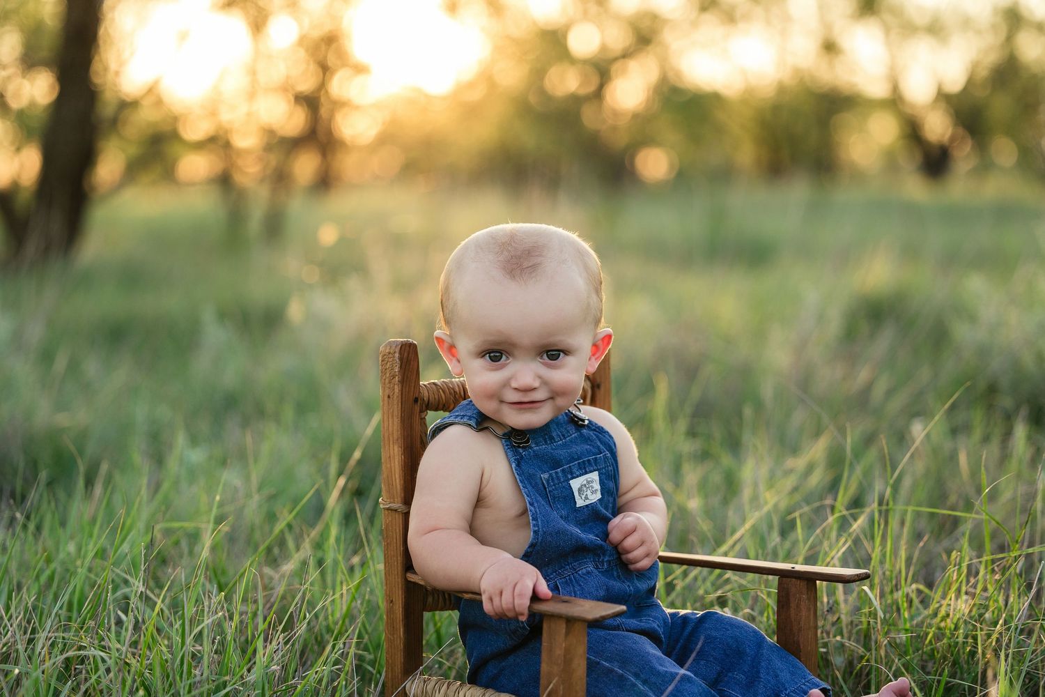 A small child in blue overalls sits on a wooden chair in a sunny grassy field during golden hour.
