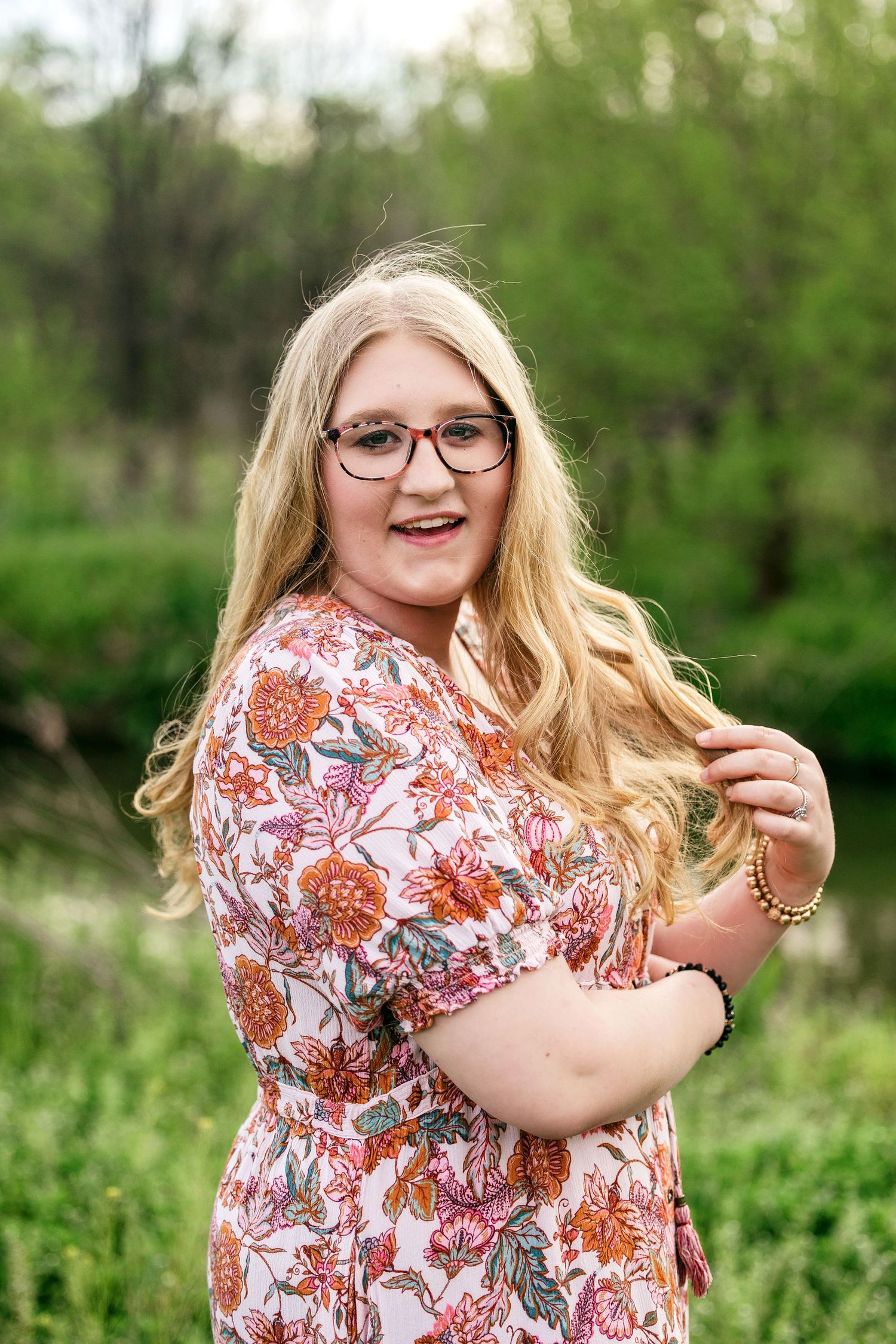 A person wearing glasses and a floral print dress poses outdoors in a natural green setting.