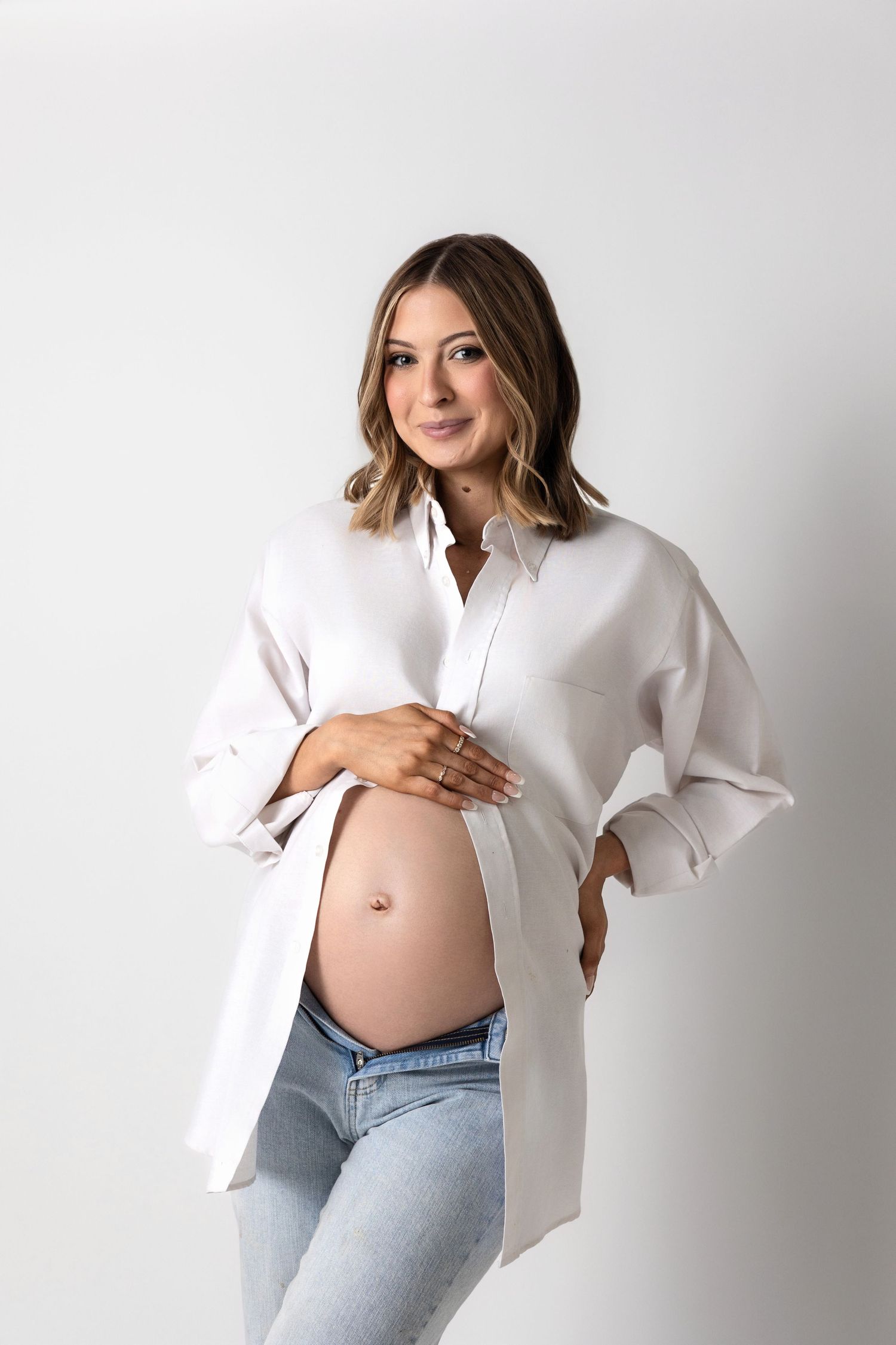 Maternity photo of woman in white button-down shirt showing baby bump against light background.