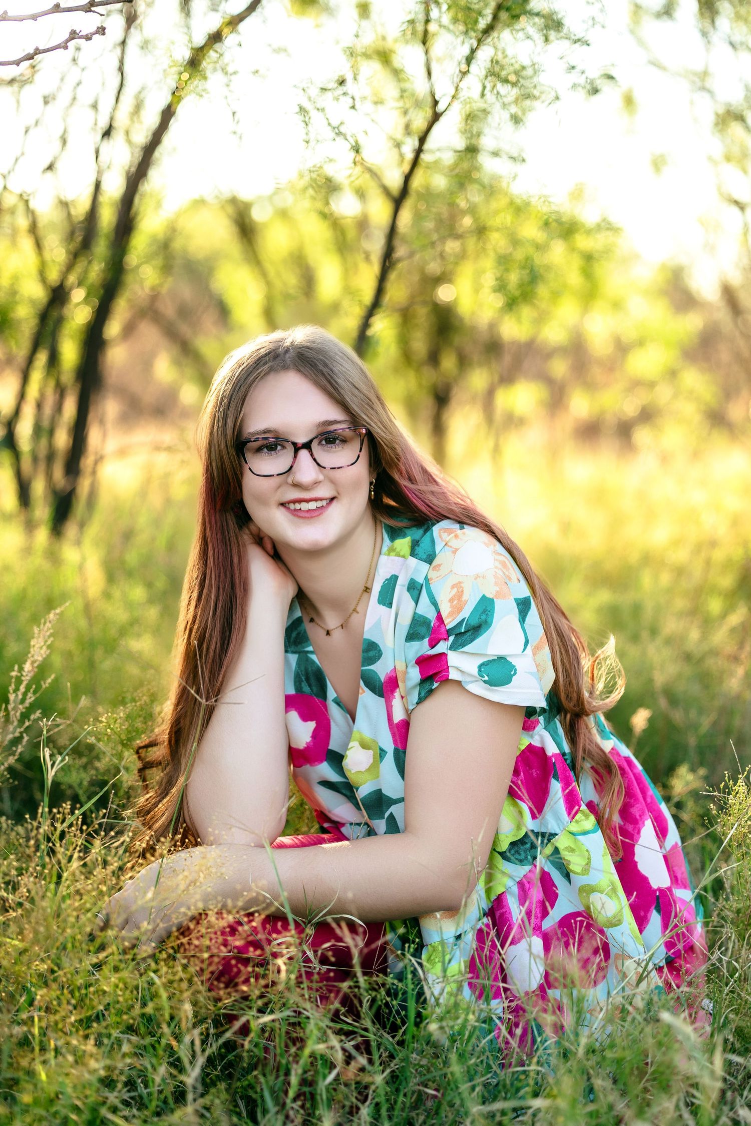 A person in glasses and a colorful floral dress poses outdoors in a sunlit meadow with tall grass and trees.