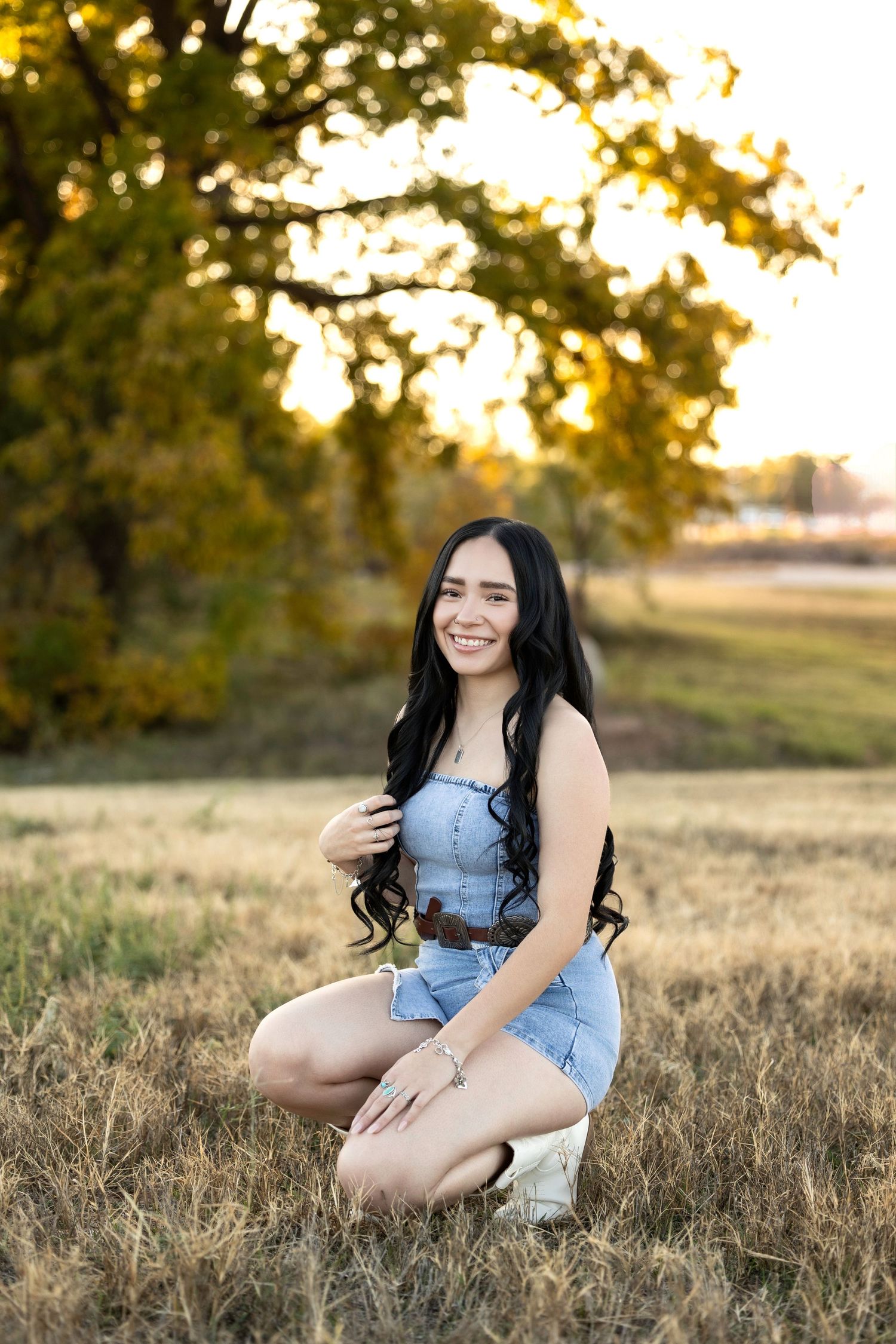 A person in denim sits in a golden field at sunset while smiling at the camera.
