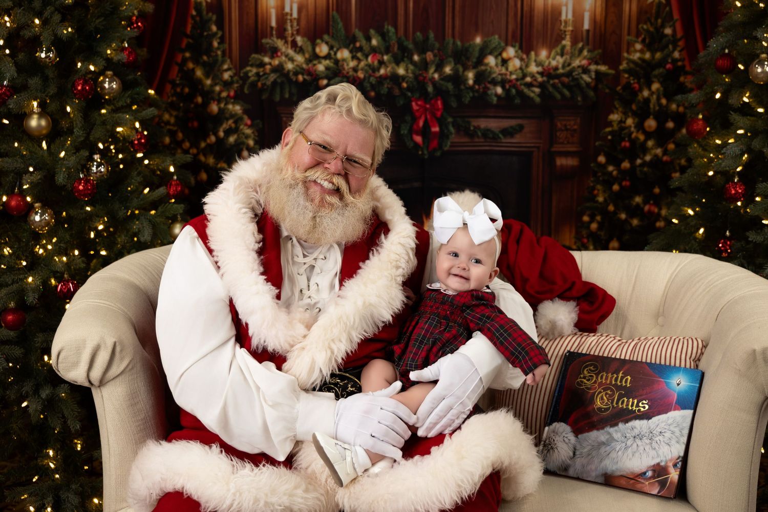 Baby in plaid dress sits on Santa Claus lap beside Christmas tree in festive holiday portrait setting.
