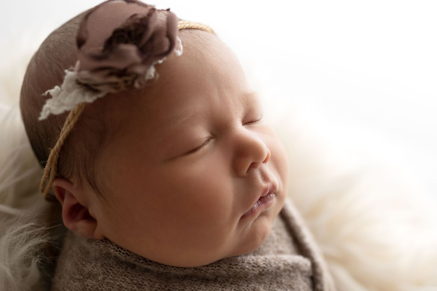 Sleeping baby wearing brown headband and gray sweater rests peacefully on soft white blanket in gentle lighting.