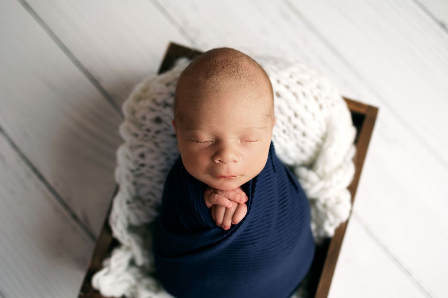 A sleeping newborn baby wrapped in a navy blue swaddle blanket rests on white textured blankets.