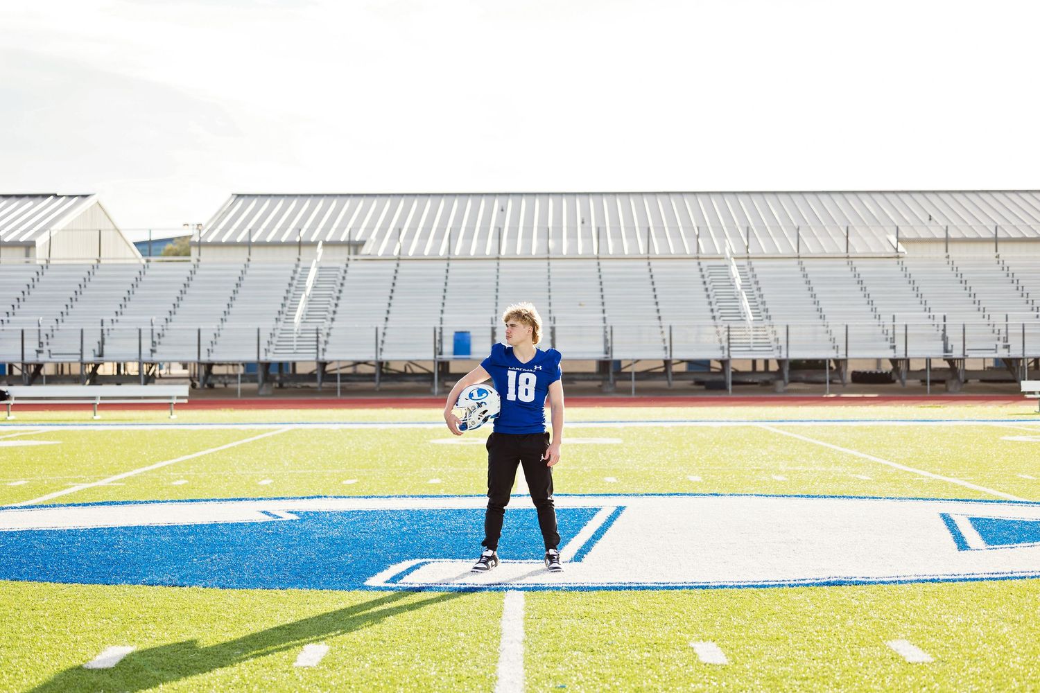 Athletes practice on a turf sports field with white line markings and greenhouses visible in the background.