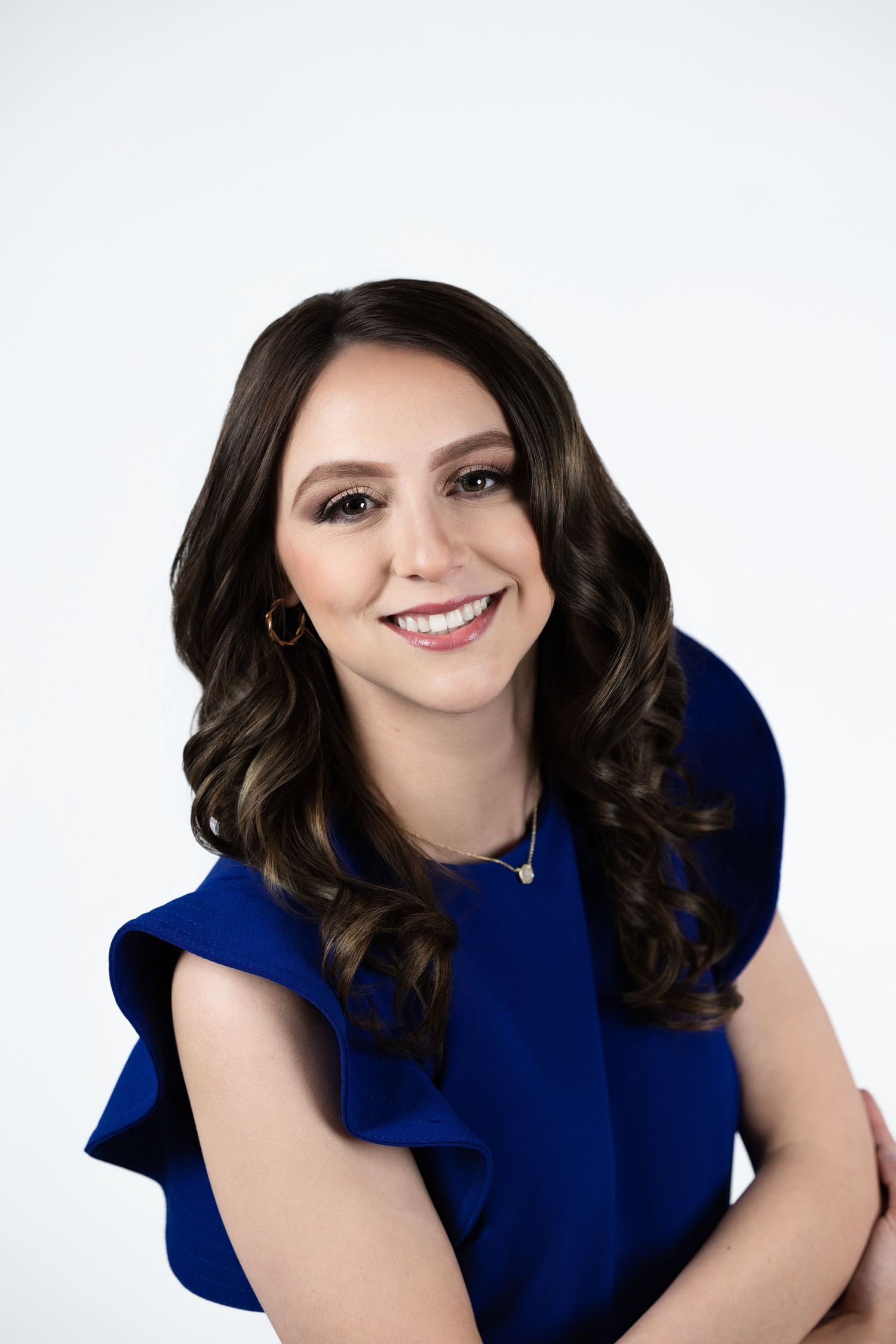 Professional portrait of someone wearing a royal blue sleeveless dress with wavy dark hair and a bright smile against white background.