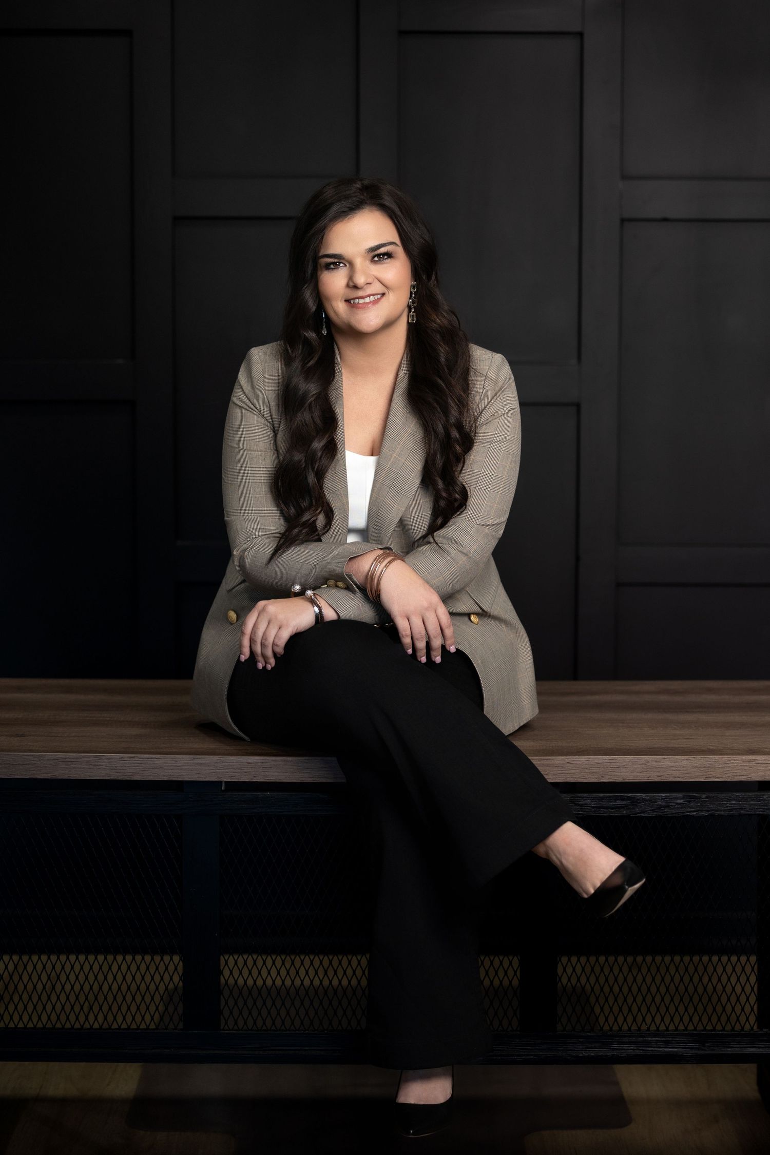 Professional studio portrait of someone in a beige blazer and black pants sitting on a wooden bench against dark background.