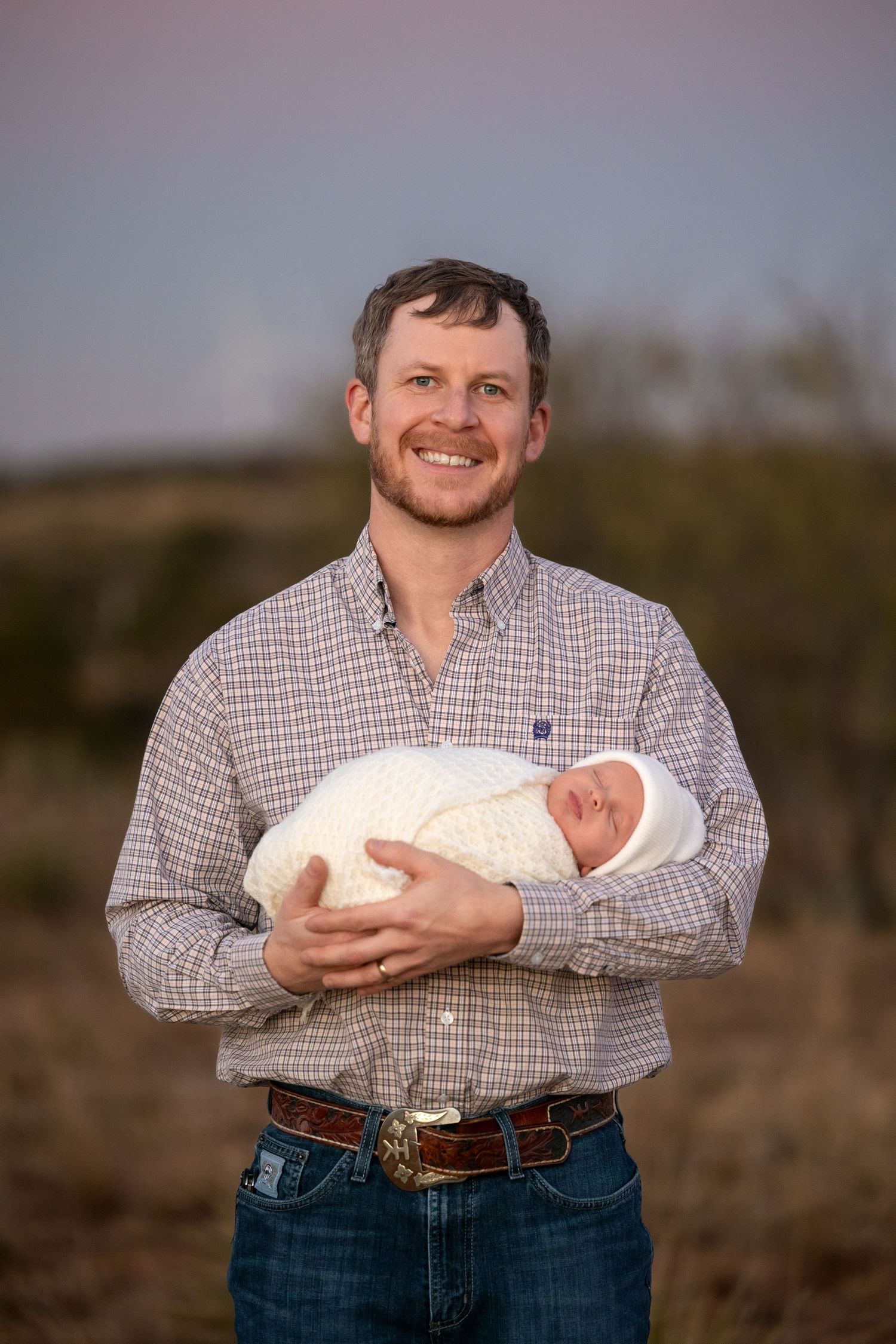 A person in a checkered shirt and jeans holds a newborn baby while standing in an outdoor setting at sunset.