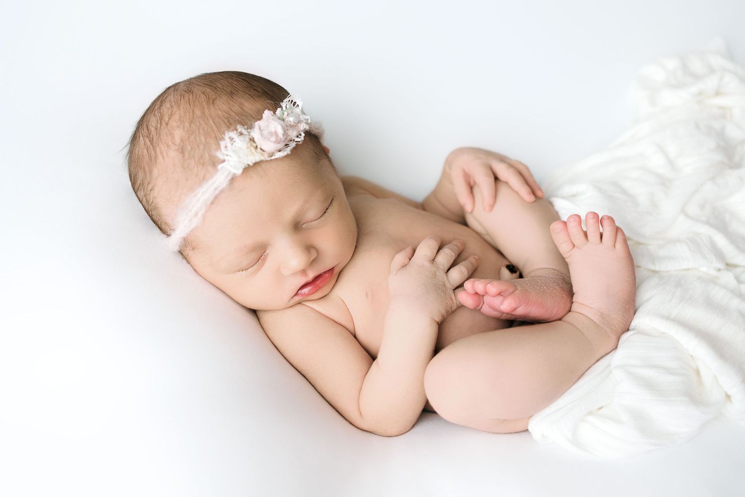 Newborn baby sleeping peacefully on white blanket wearing delicate lace headband in soft natural light.