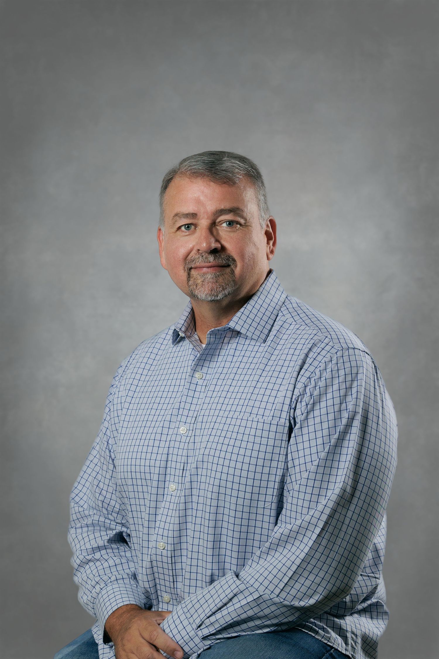 Professional studio portrait of someone wearing a light blue checkered button-down shirt against a grey backdrop.
