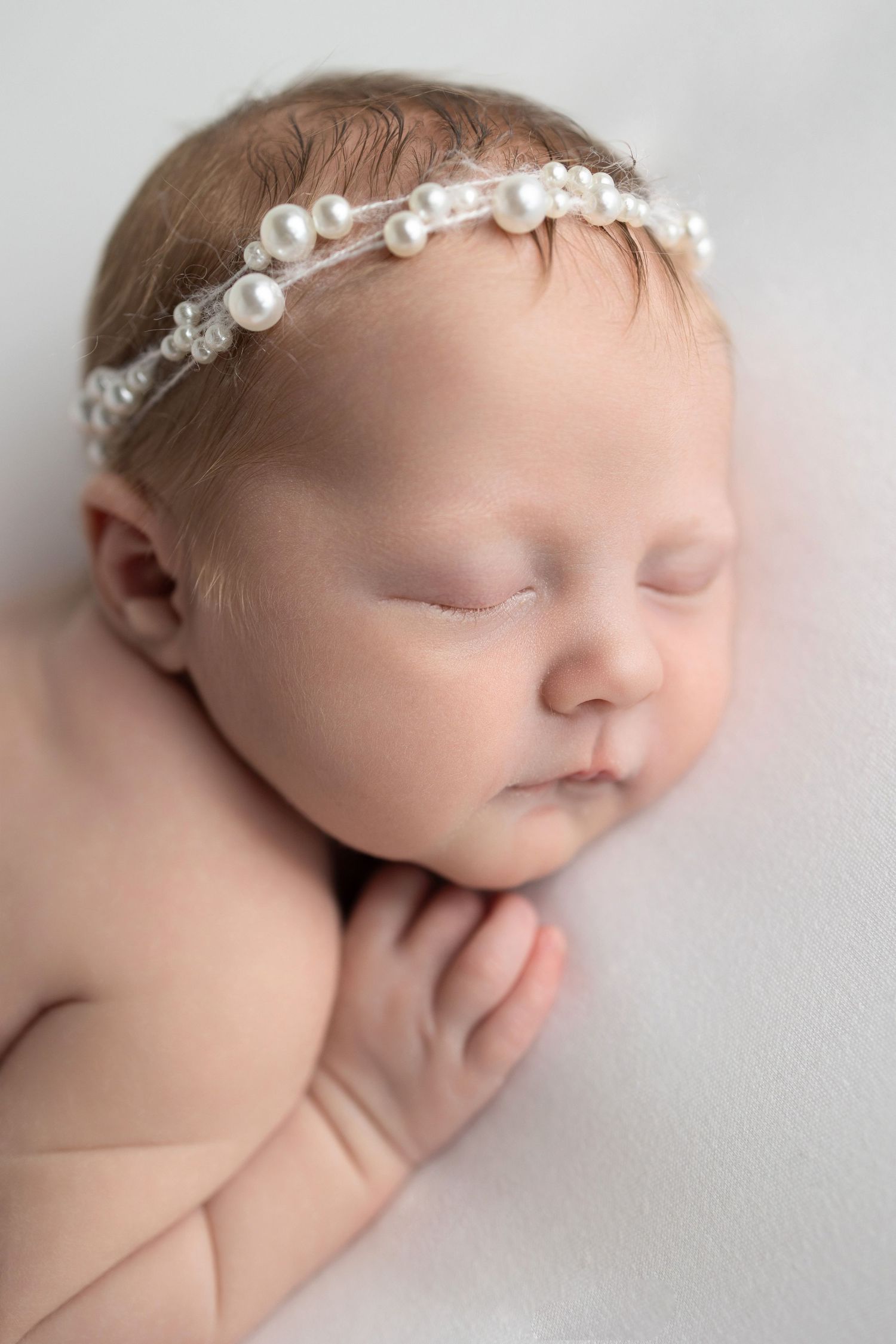 A peaceful sleeping newborn wearing a delicate pearl headband rests against a soft white backdrop.