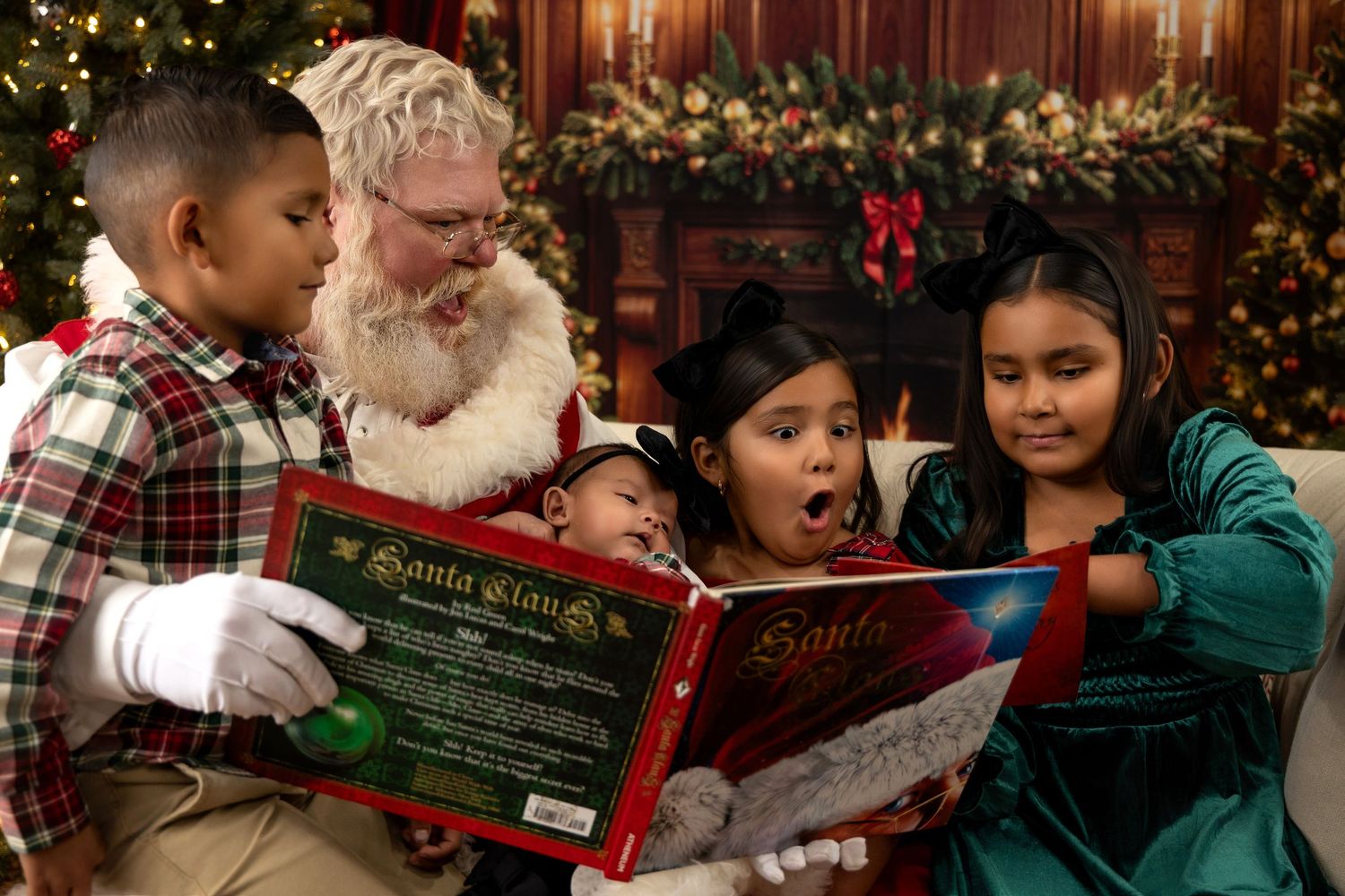 Santa Claus reads a festive book with three excited children by a decorated Christmas fireplace.