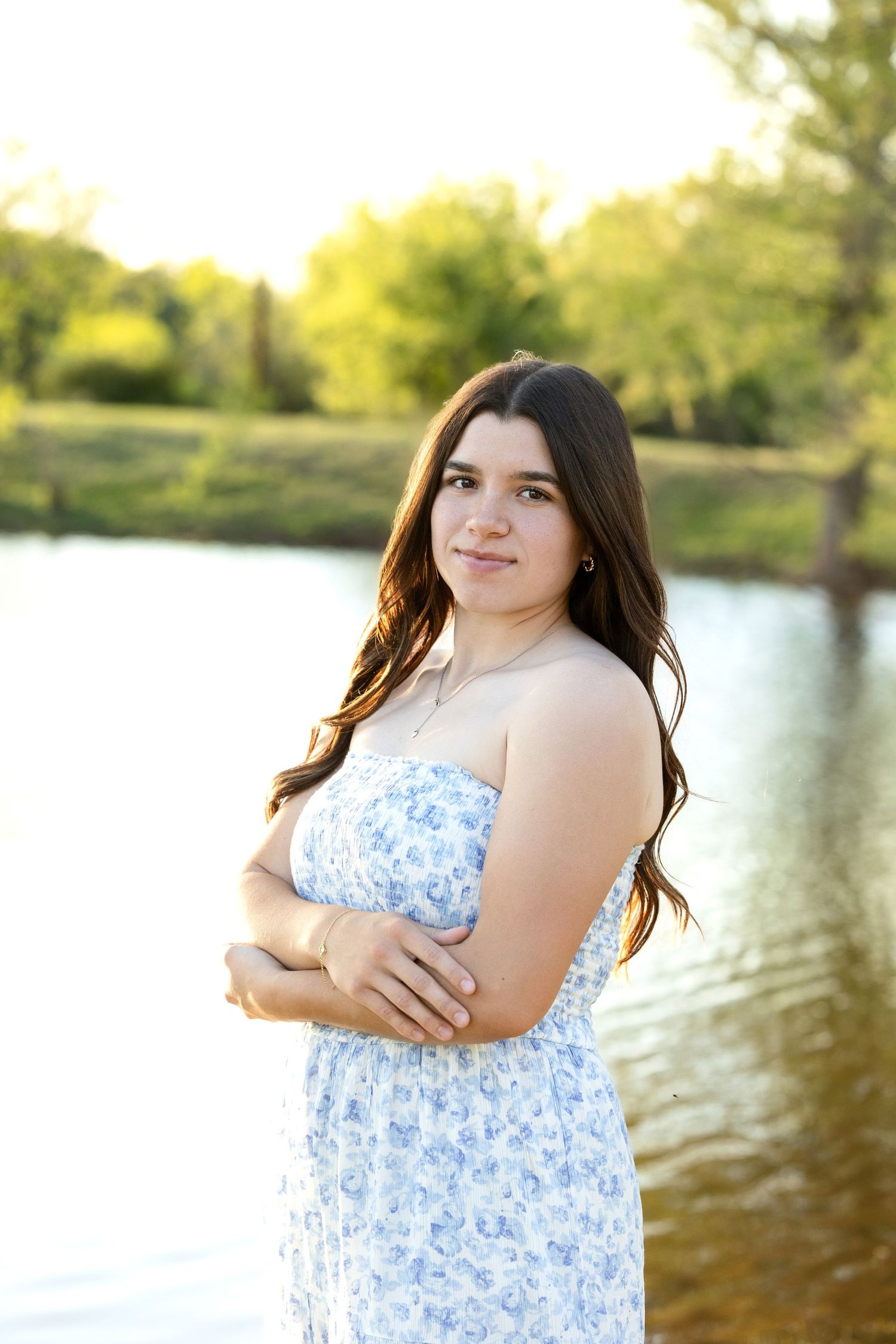 A young woman in a blue and white floral dress poses by a lake at sunset with trees in the background.