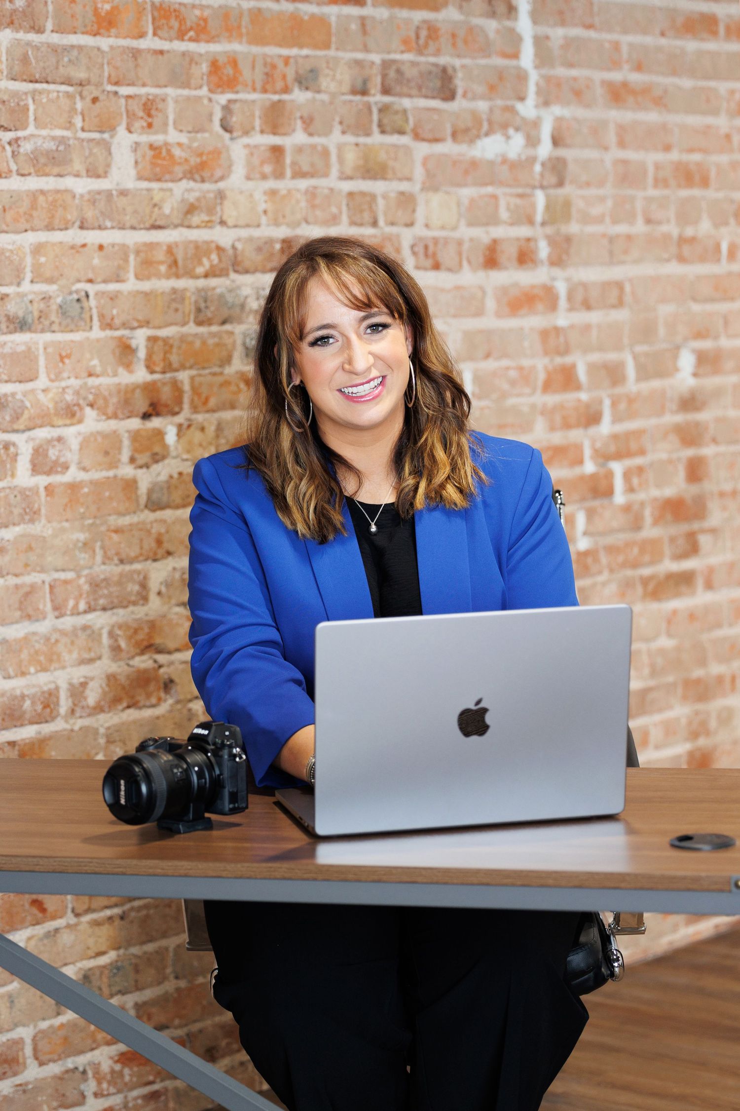 Professional photographer sits at desk with MacBook laptop and DSLR camera against exposed brick wall background.
