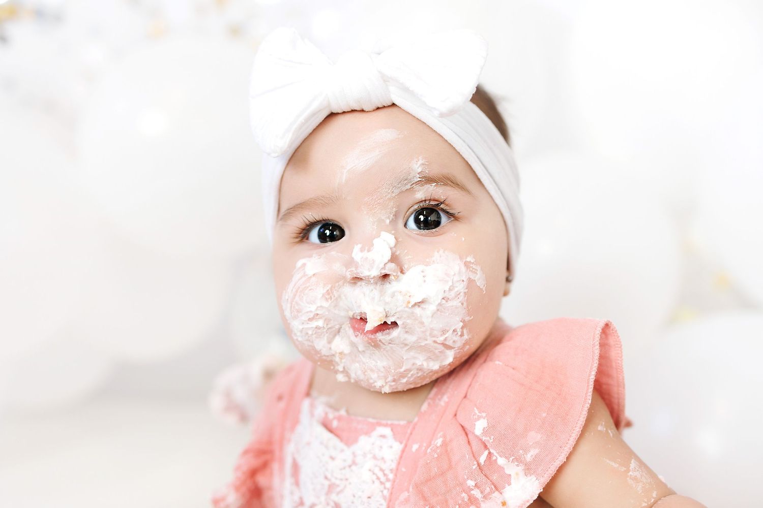 Baby wearing pink bib and white headband with messy face covered in yogurt or cream during mealtime.