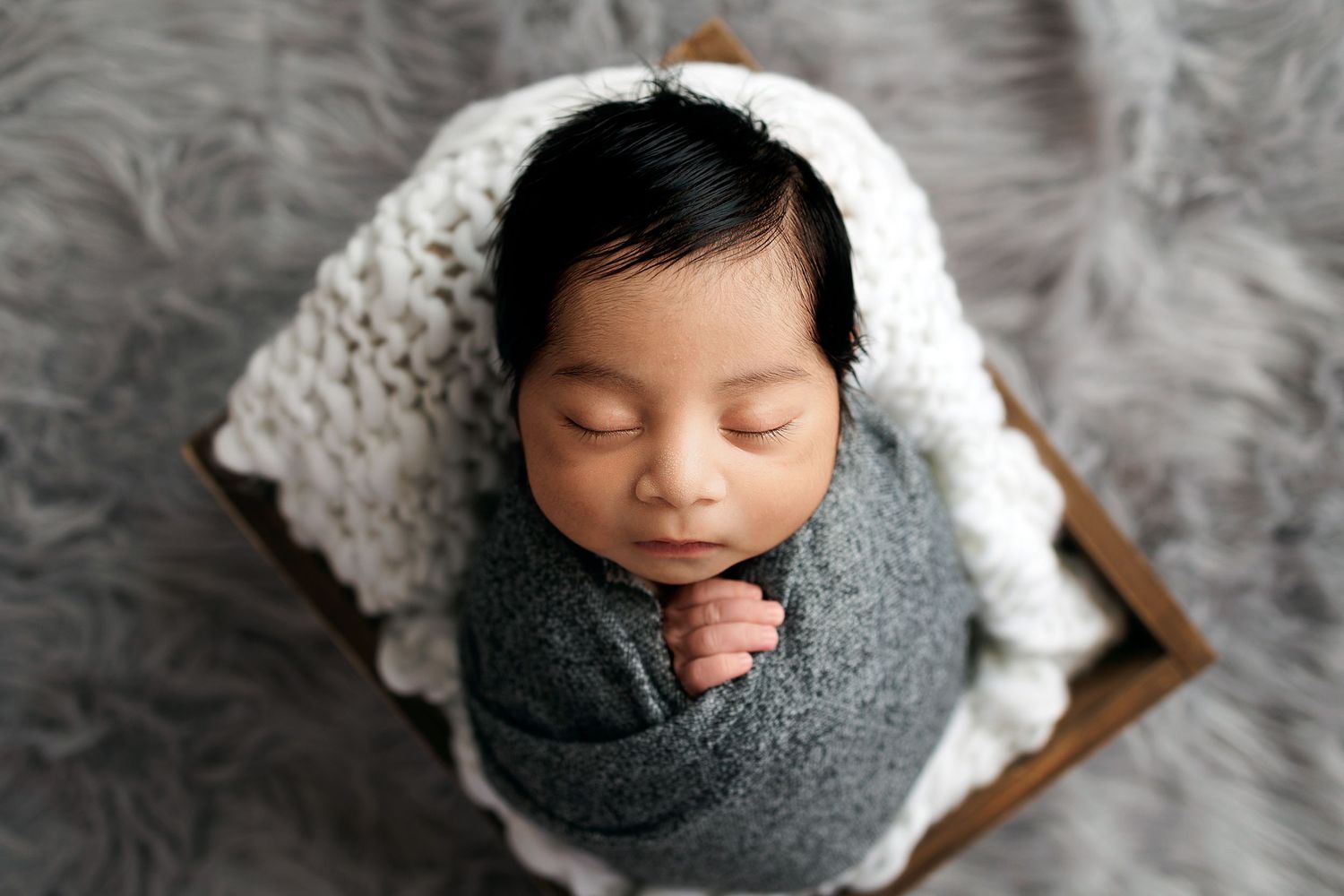 Overhead view of sleeping newborn wrapped in gray swaddle on white blanket and gray fur background.