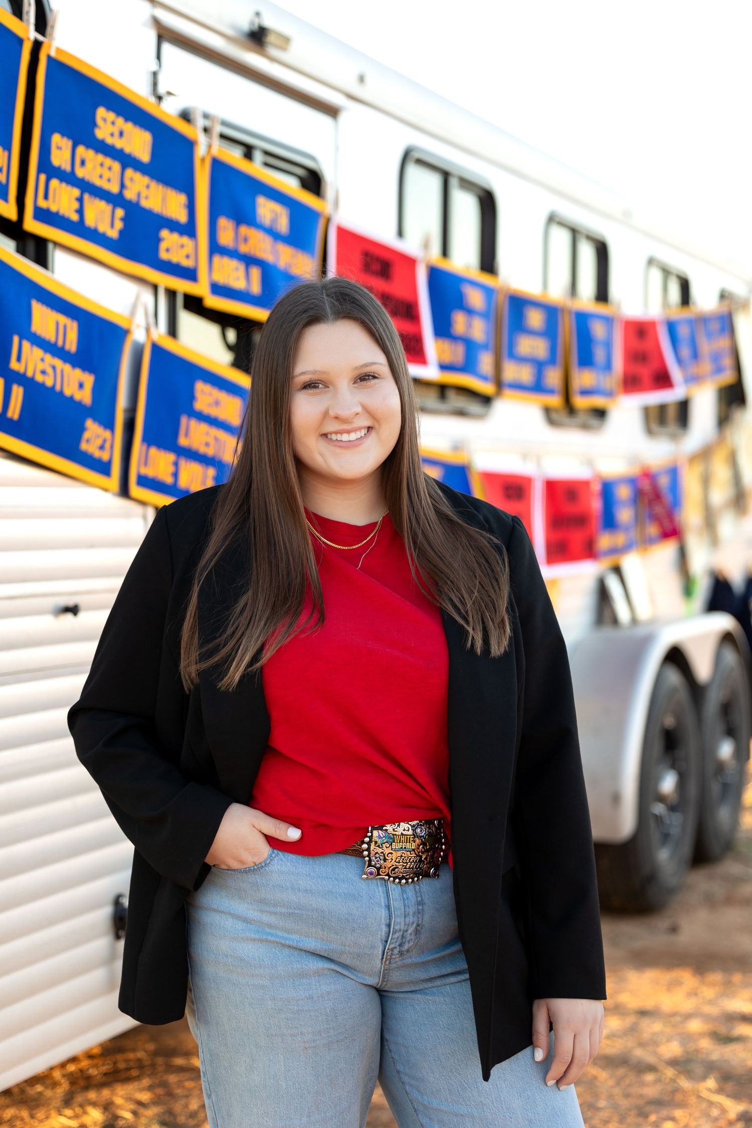 A person in a red top and black cardigan stands smiling in front of a livestock show trailer with banners.