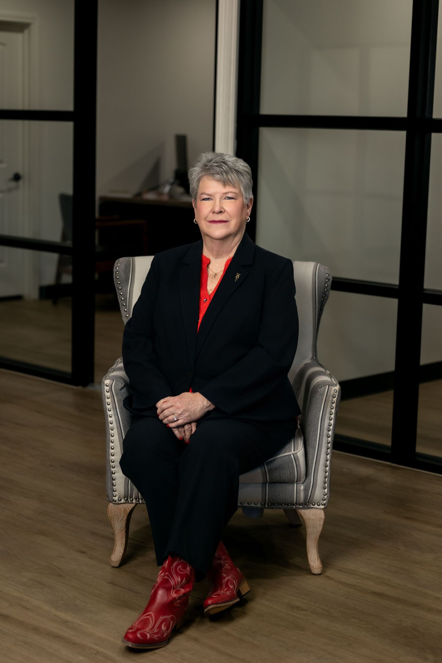 Professional seated in a gray armchair wearing a black suit and red boots against a modern office interior.