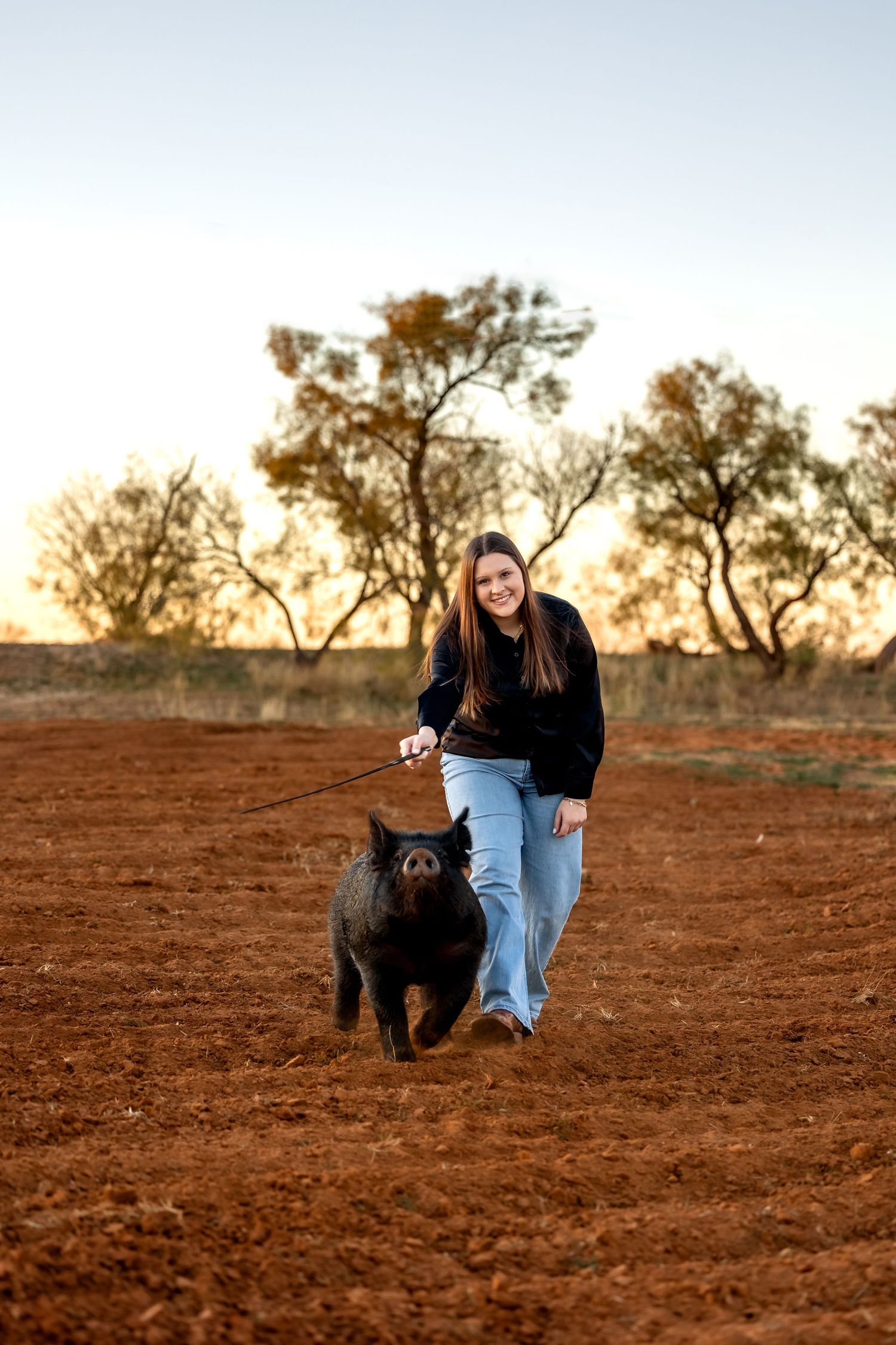 A person and black dog sit together on red desert soil at sunset with trees silhouetted in the background.