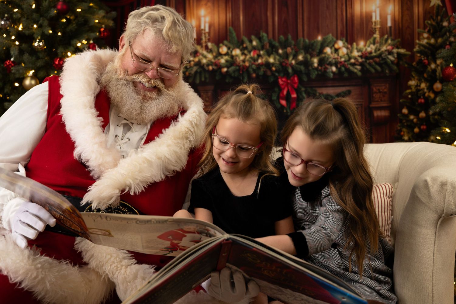 Santa Claus reads a festive storybook with two young girls in glasses beside a decorated Christmas tree.