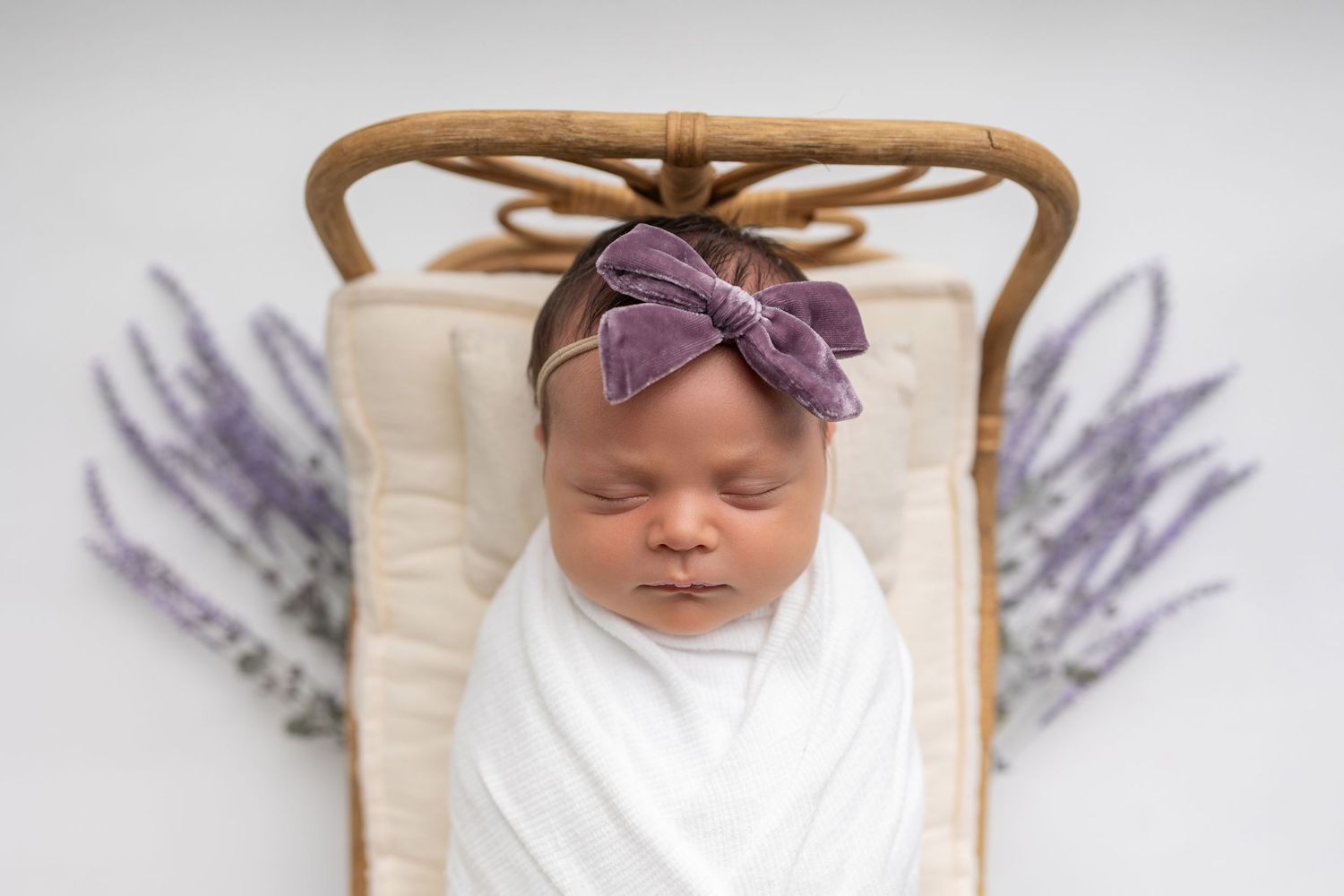 A baby in white swaddle blanket lies in wooden basket with lavender sprigs on either side.
