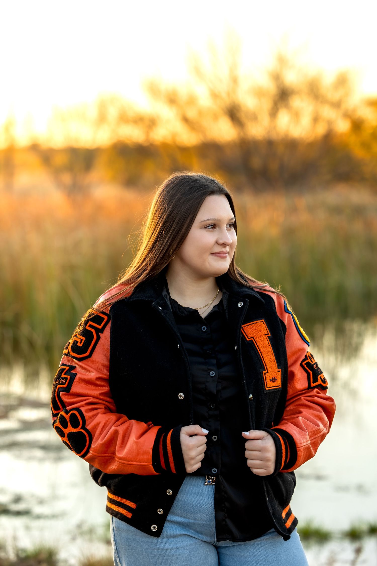A person in a black and orange varsity jacket poses outdoors during golden hour by a lake.