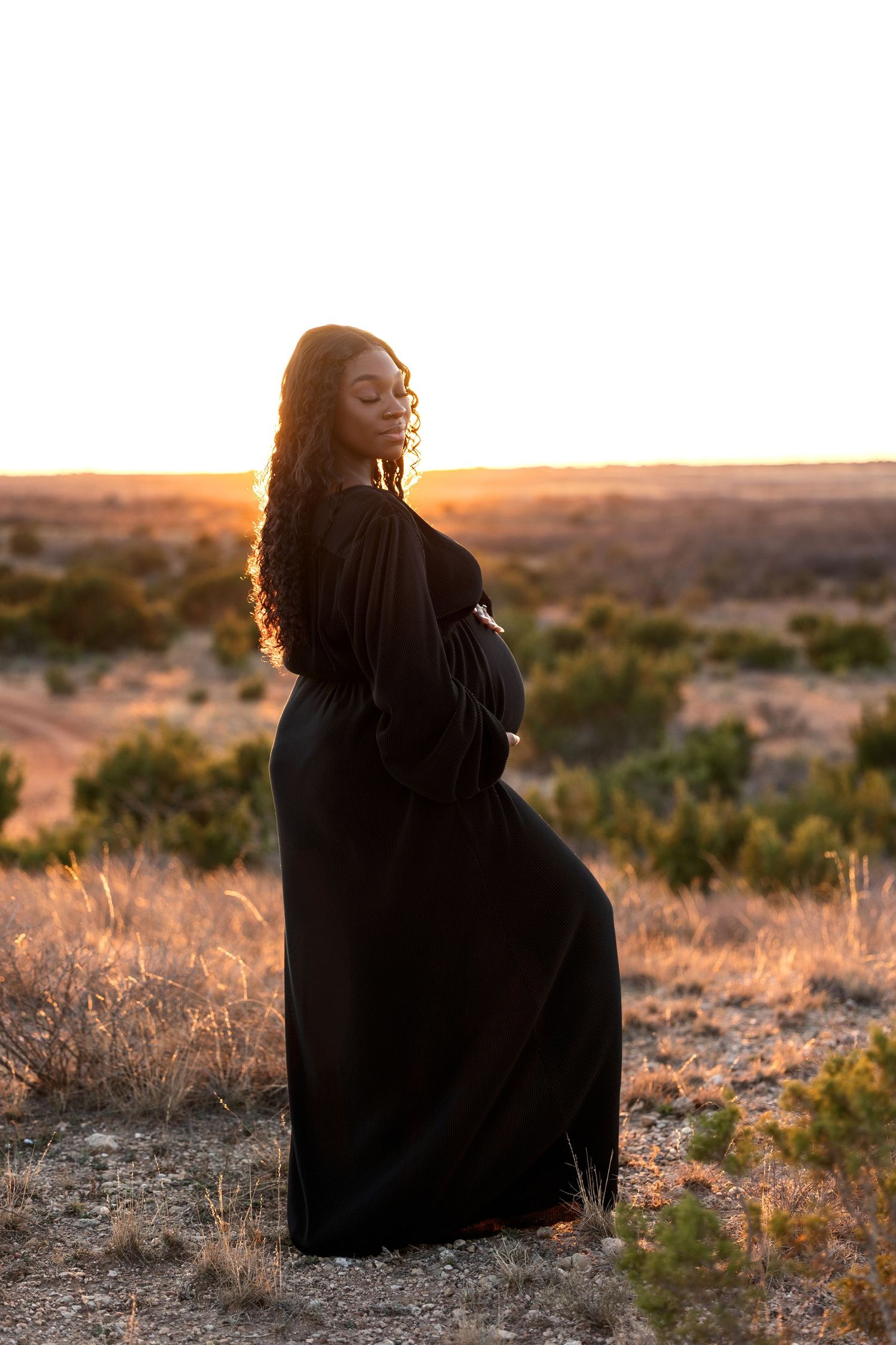 A person in a black dress sits in a desert landscape at golden hour as sunlight creates a warm atmospheric glow.