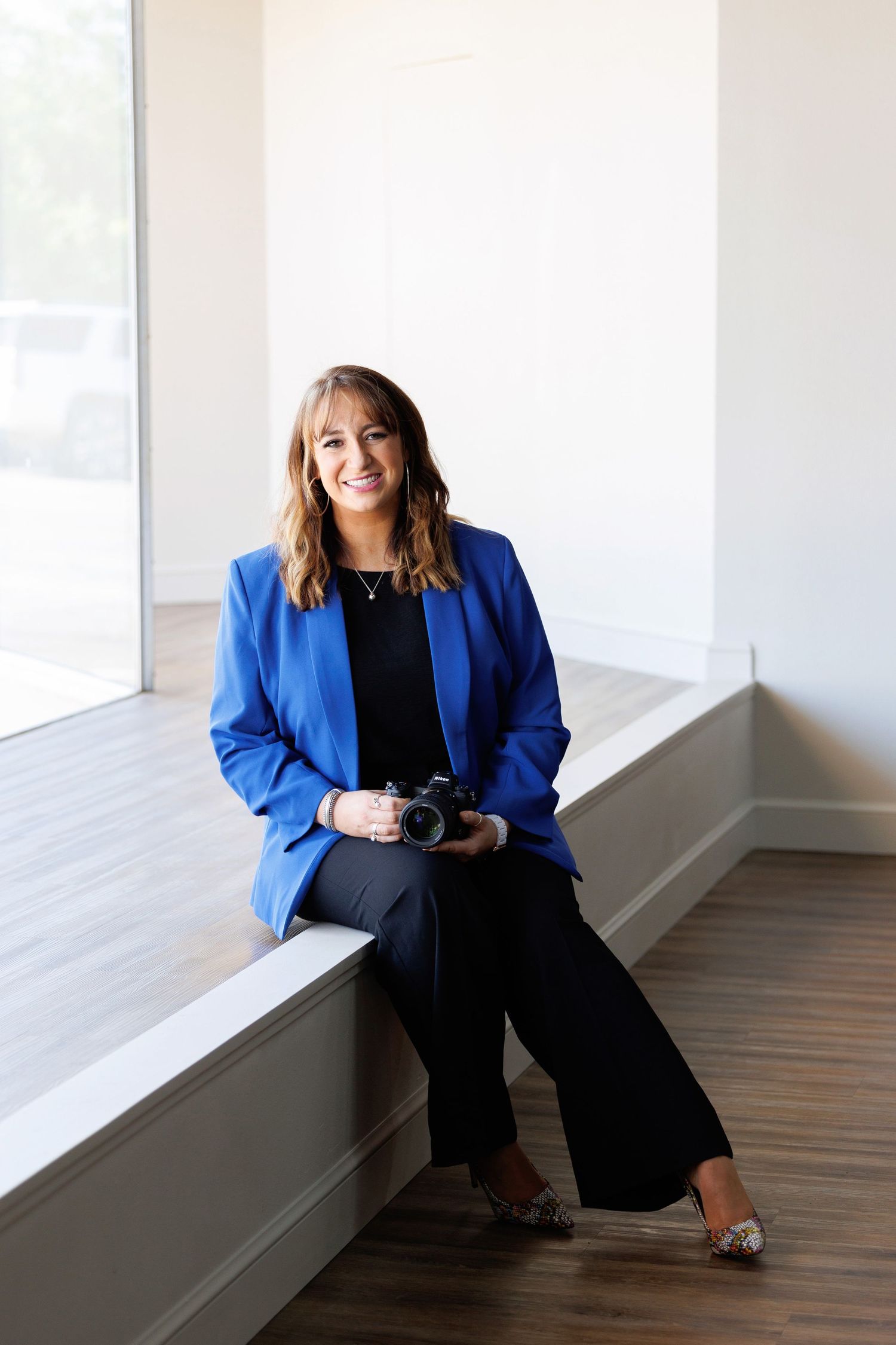 Professional wearing a royal blue blazer and black pants sits casually by a window in a modern interior space.