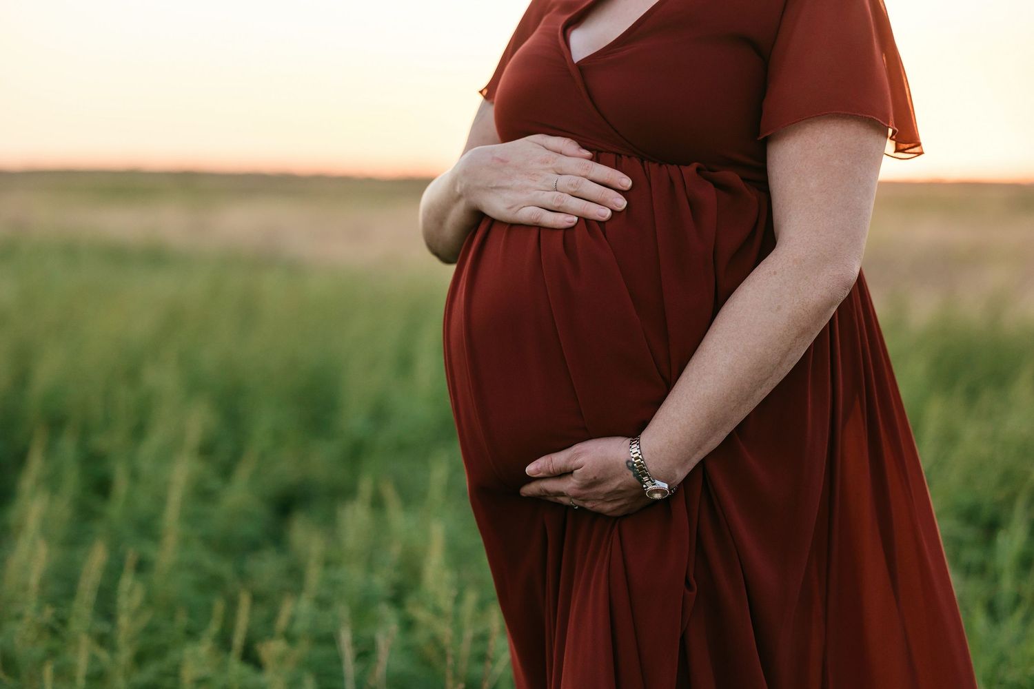 A woman in a burgundy dress cradles her pregnant belly while standing in a lush green field at sunset.