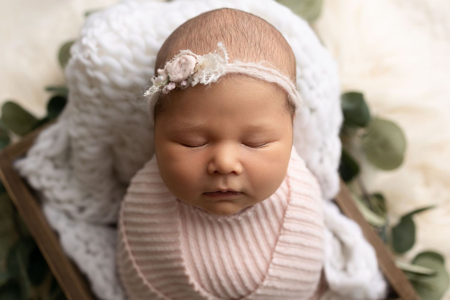 Sleeping newborn baby wearing pink knit clothing and headband surrounded by white blankets and eucalyptus leaves.