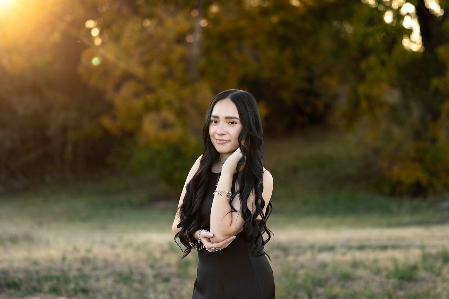 A person with long dark hair poses outdoors at sunset wearing a black dress in a golden-lit field.