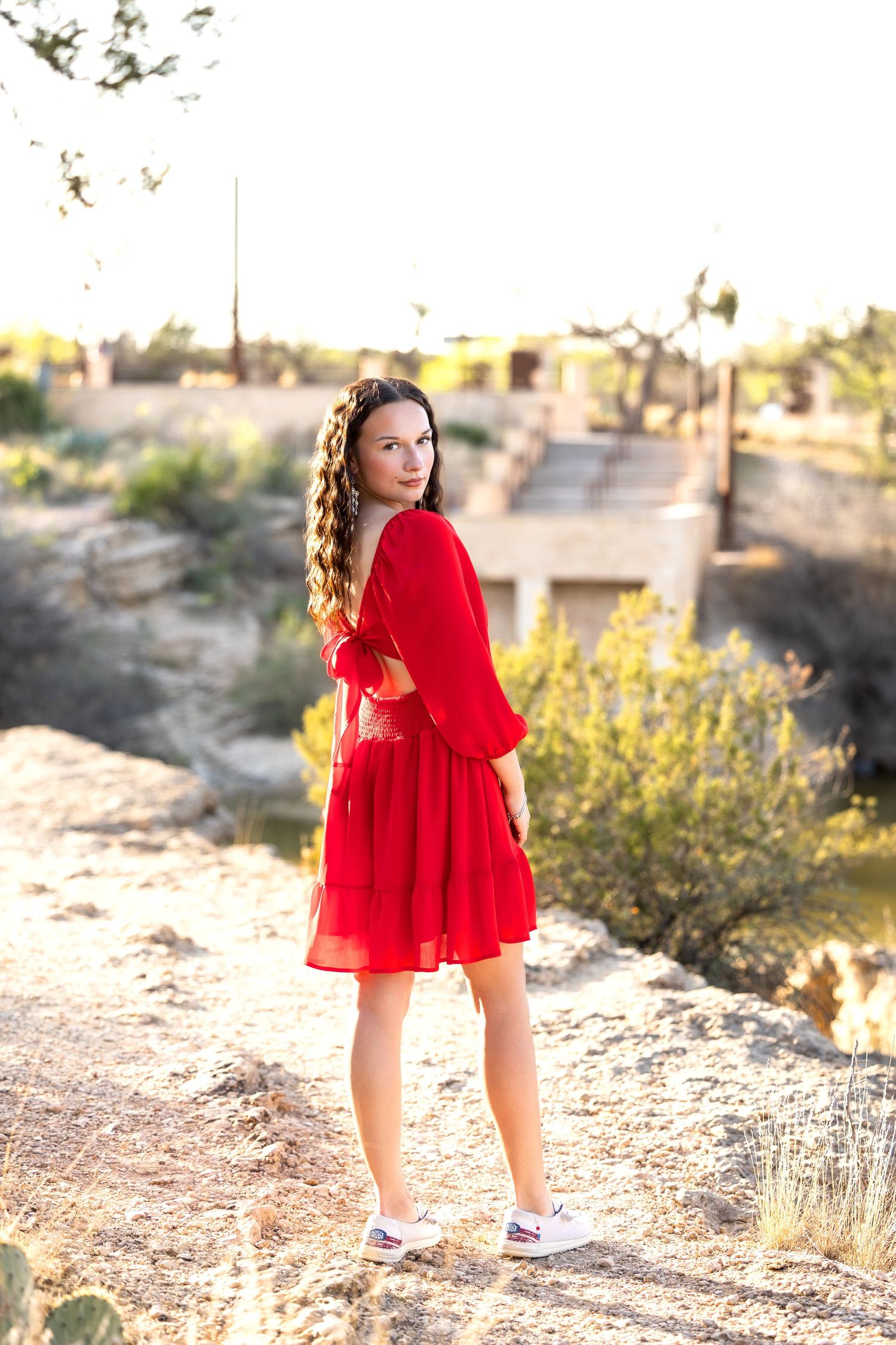 A person in a bright red flowy dress poses against a desert landscape with yellow wildflowers at sunset.