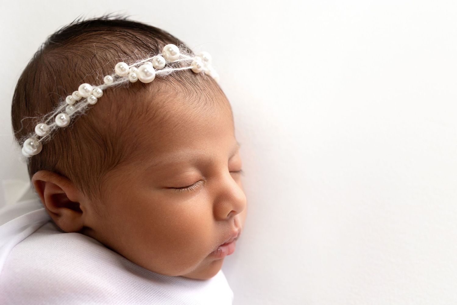 A sleeping newborn baby wearing a delicate white lace headband rests peacefully on soft white bedding.