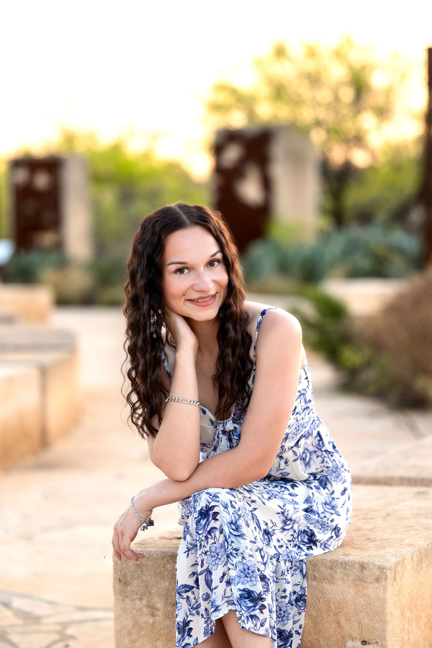 A woman in a blue and white floral dress sits on a stone wall in a garden setting during golden hour.