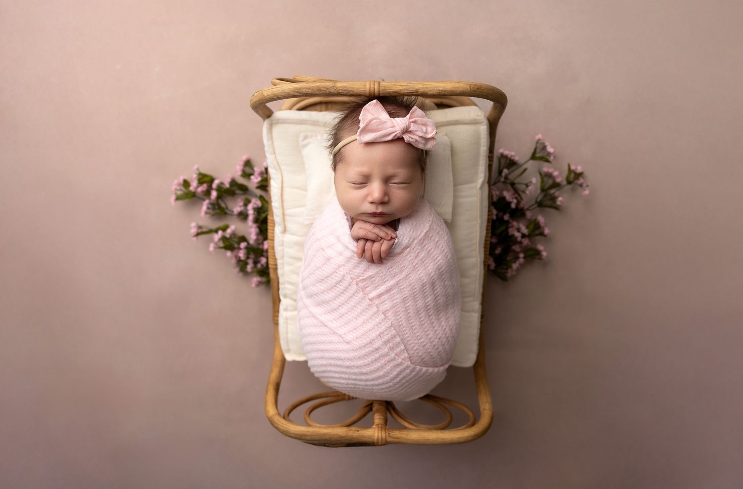 A sleeping newborn wrapped in pink swaddle lies in a vintage wooden basket with floral decorations on either side.