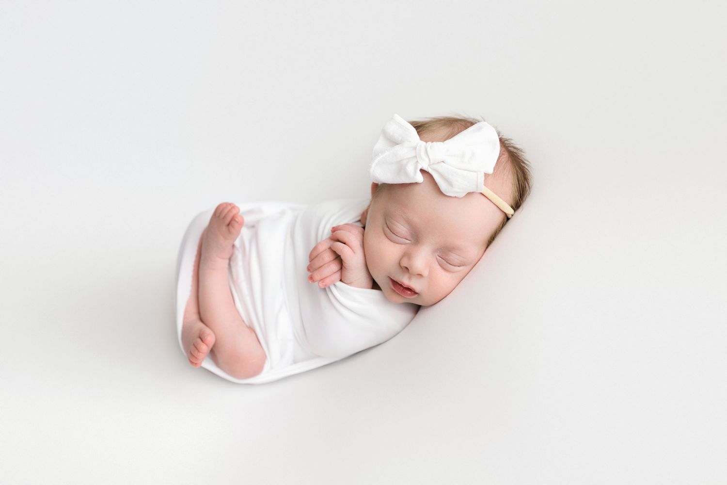Sleeping newborn swaddled in white fabric with a white bow headband on light background.