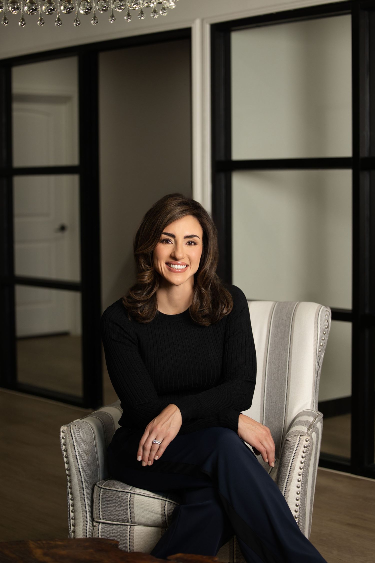 Professional portrait of someone in a black sweater seated in an elegant gray armchair against glass doors.