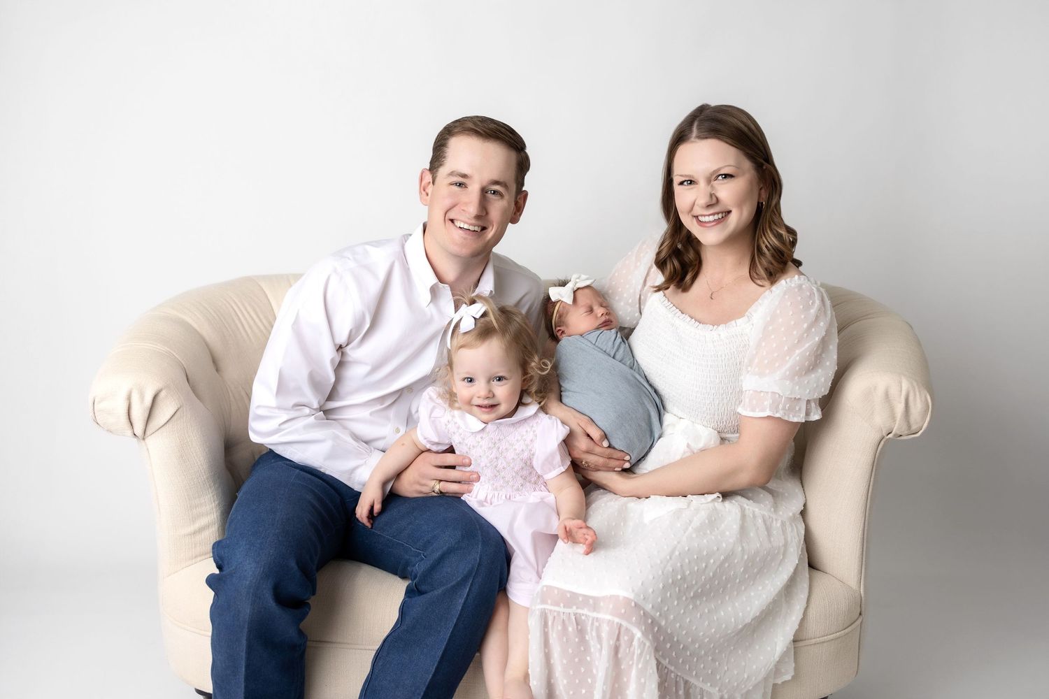 Family wearing coordinated white and beige outfits poses together on cream colored couch in studio setting.