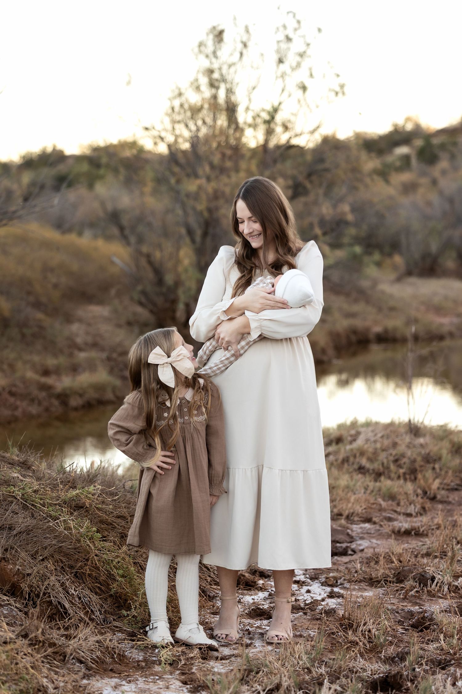 Two figures in contrasting brown and white outfits share tender moments by a serene lake at sunset.